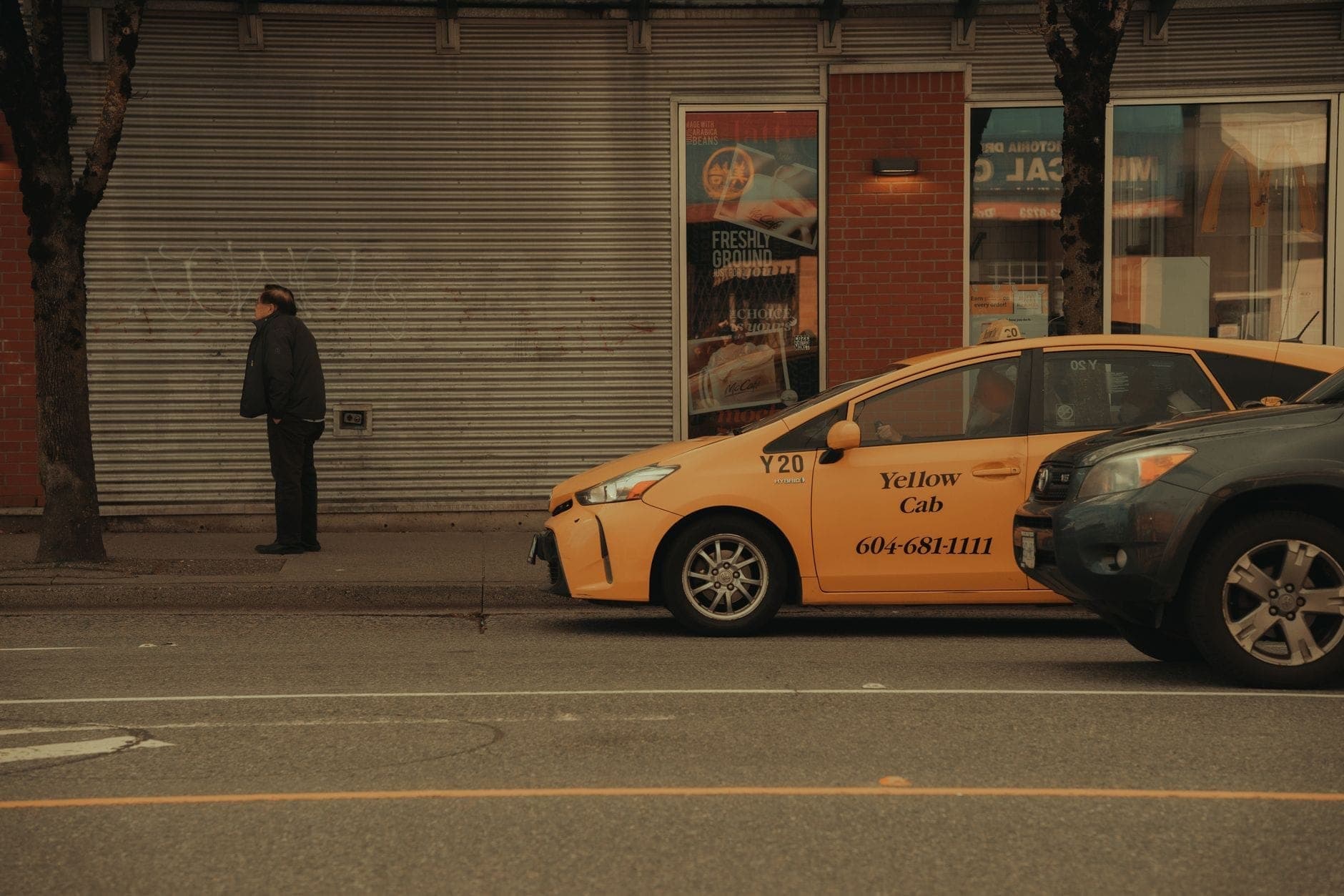 แท็กซี่ Yellow Cab จอดอยู่บนถนนในเมือง Vancouver ช่วงกลางวัน มีคนเดินเท้าอยู่ใกล้ๆ พร้อมด้วยร้านค้าและต้นไม้ริมทางเท้า