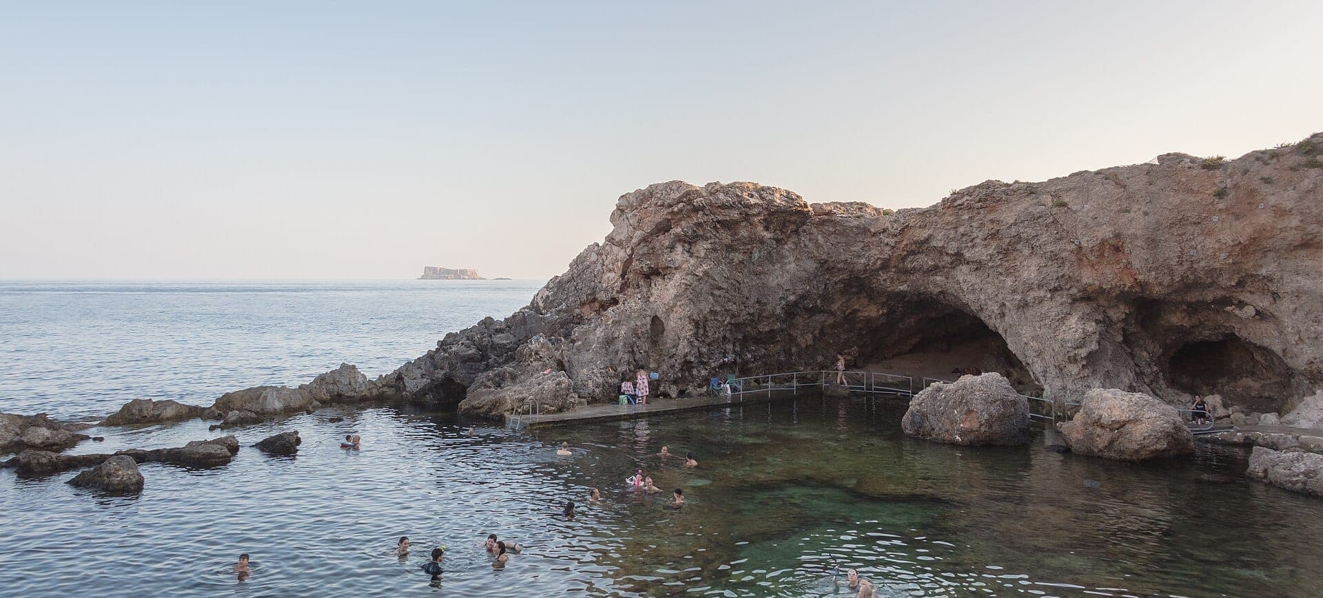 Klares Meerwasser mit Schwimmern nahe dem felsigen Eingang der Għar Lapsi-Höhle, umgeben von schroffen Klippen an der Südküste Maltas.