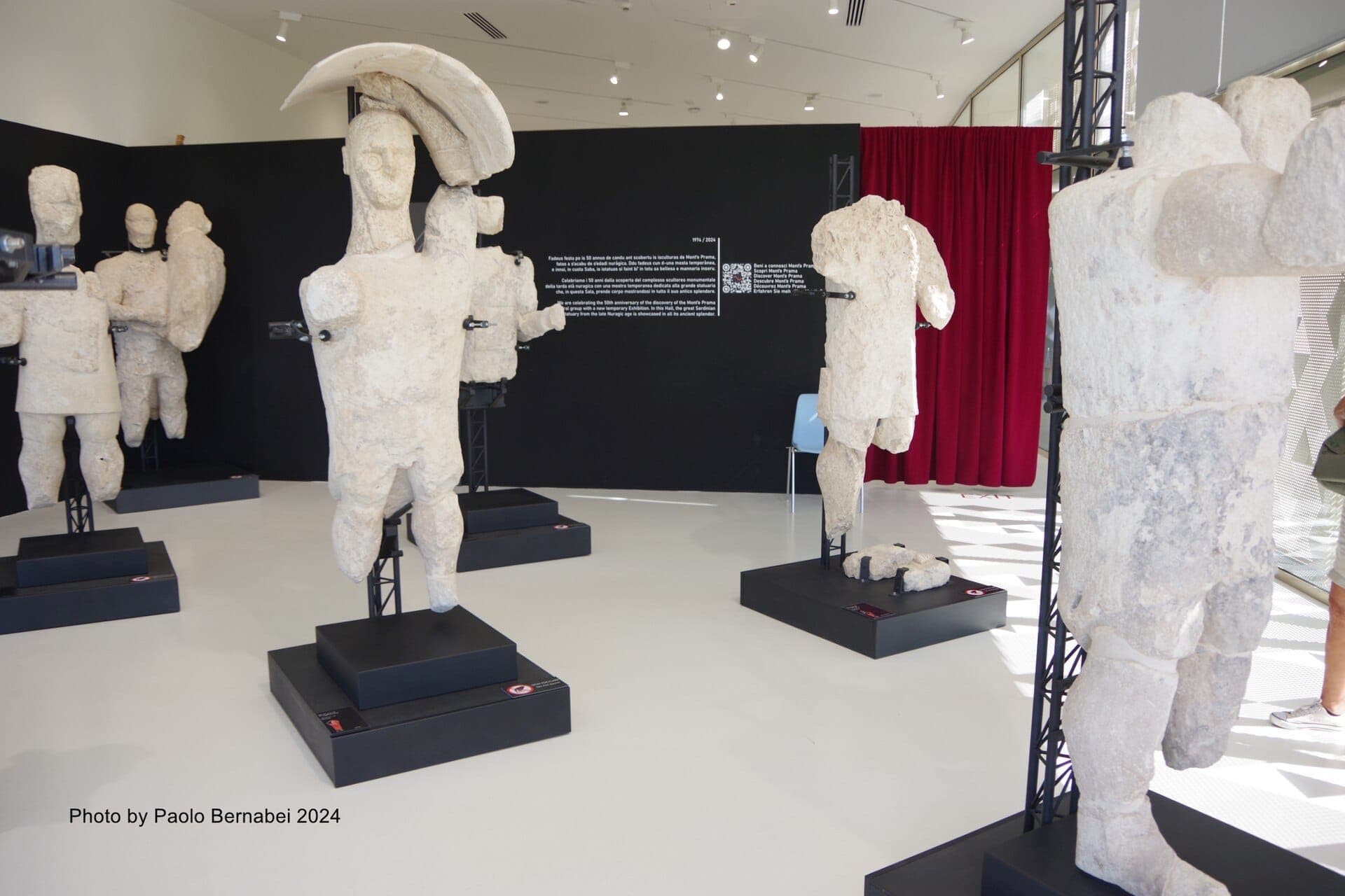 Varias antiguas estatuas de piedra de los Gigantes de Mont'e Prama expuestas sobre pedestales negros en una sala de exposición moderna y luminosa.