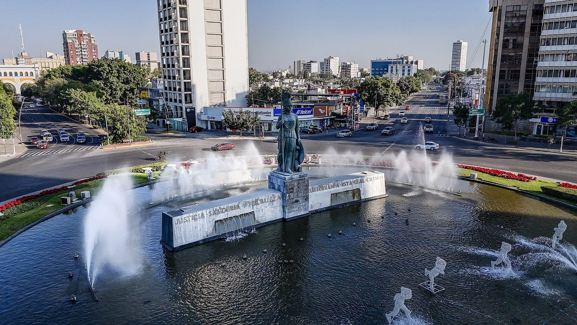 ภาพมุมสูงของ Glorieta de La Minerva พร้อมรูปปั้นบรอนซ์ที่โดดเด่น น้ำพุโดยรอบ ถนนในเมือง และอาคารสูงในวันที่อากาศแจ่มใส