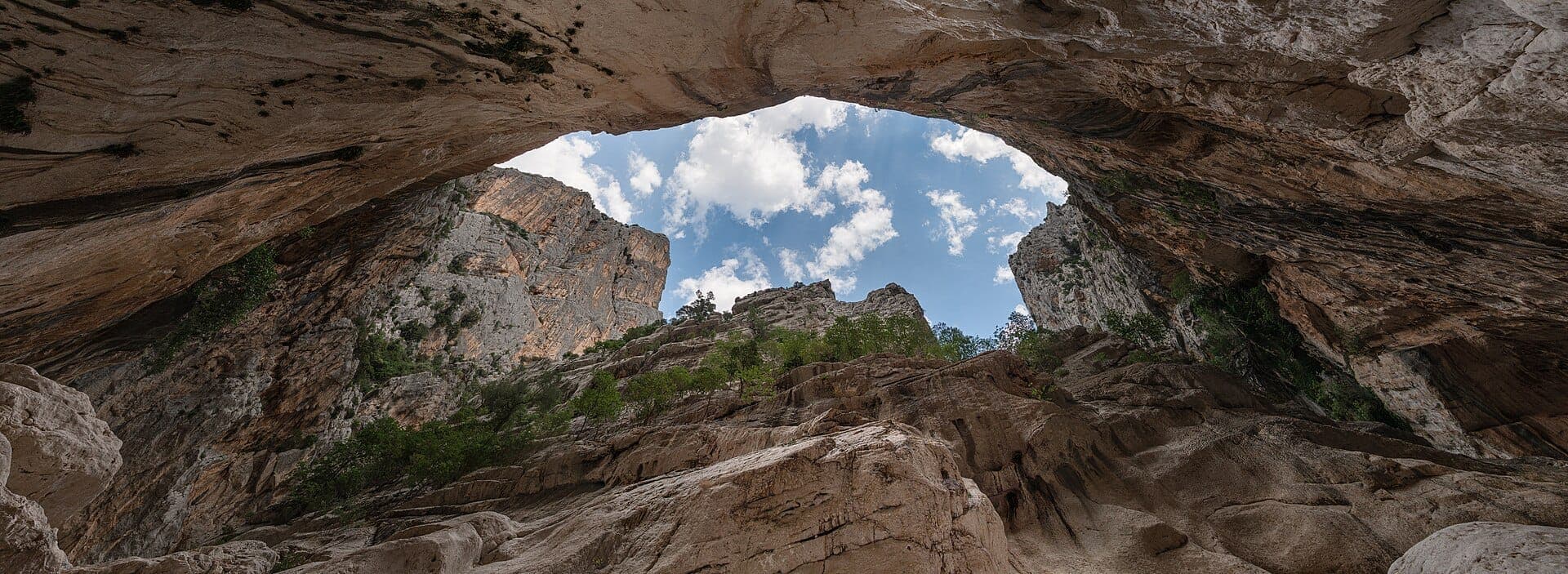 Vista gran angular desde el interior de Gola di Su Gorropu que muestra las imponentes paredes del cañón, terreno rocoso, arbustos verdes y un cielo azul con nubes dispersas.