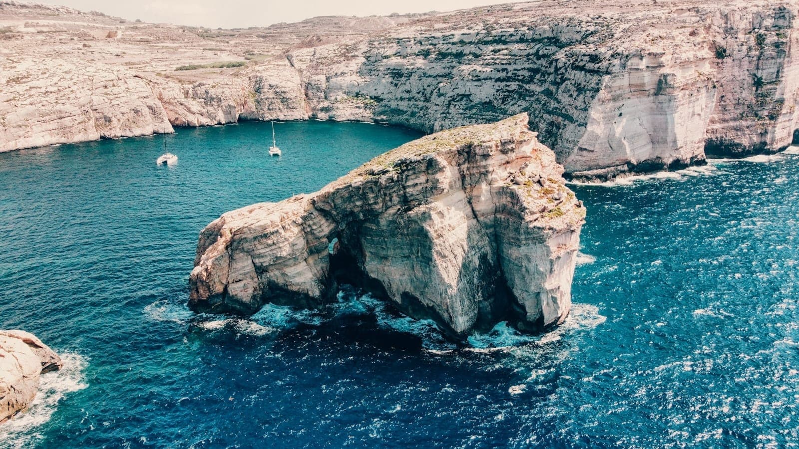 Dramatische Küstenklippen und eine große Felsformation, umgeben von türkisblauem Wasser in Gozo, Malta – an einem sonnigen Tag aus der Vogelperspektive aufgenommen.