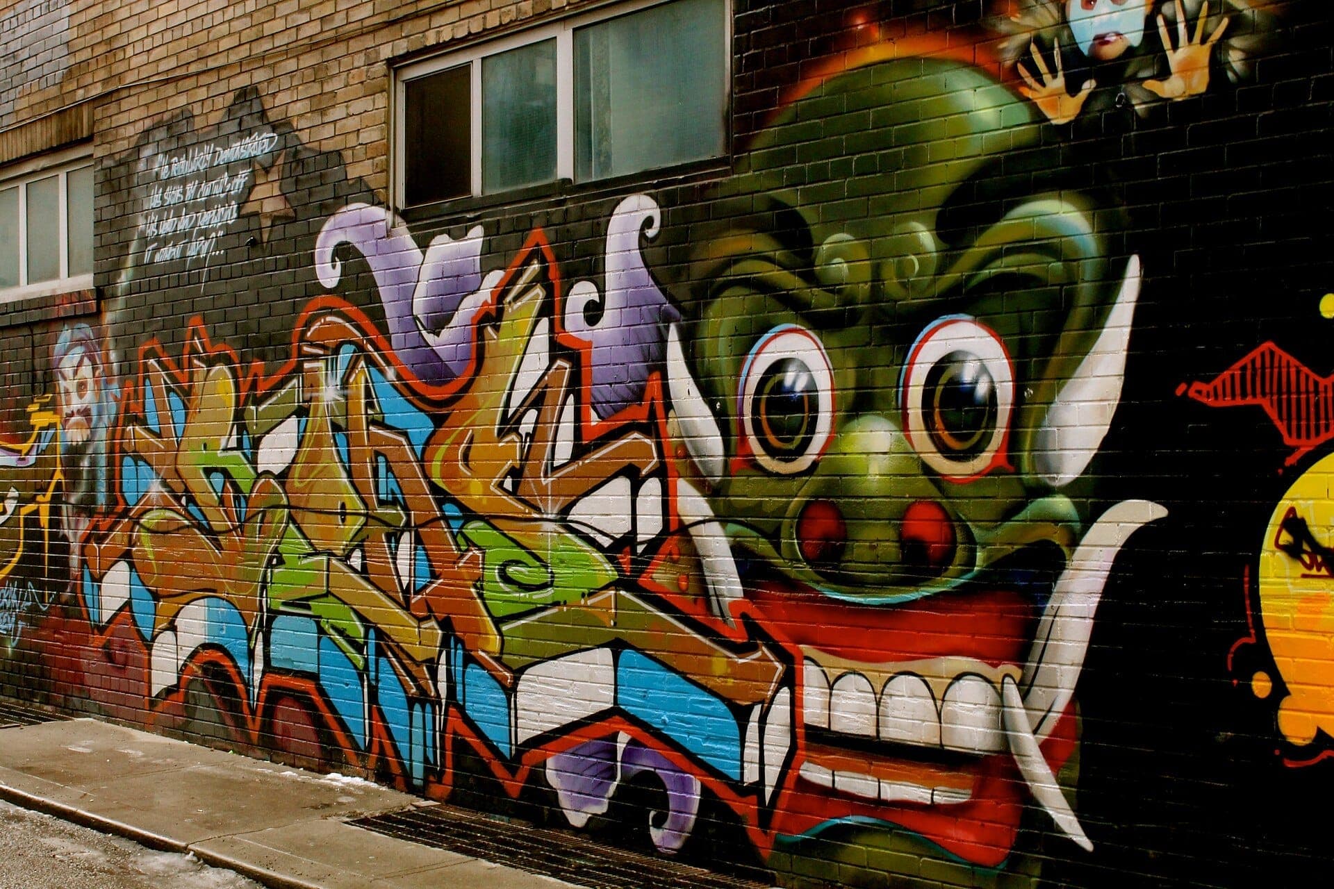 Arte di strada colorata e un grande murale con un volto di mostro verde su un muro di mattoni nella Graffiti Alley di Toronto, con tag spray e contesto urbano.