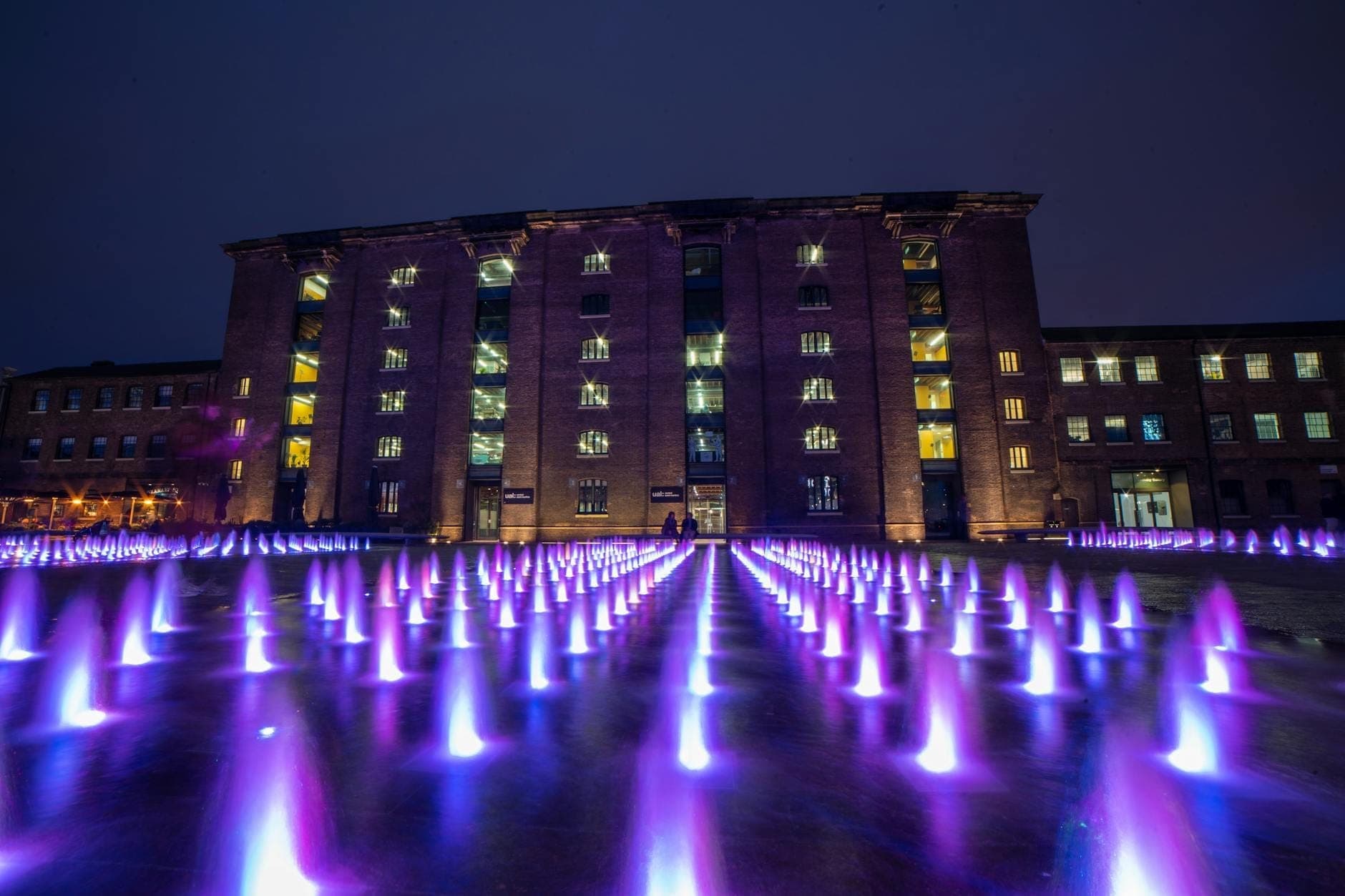 Pemandangan malam Granary Square dengan air mancur bercahaya ungu di depan Gedung Granary bersejarah di King's Cross, London.