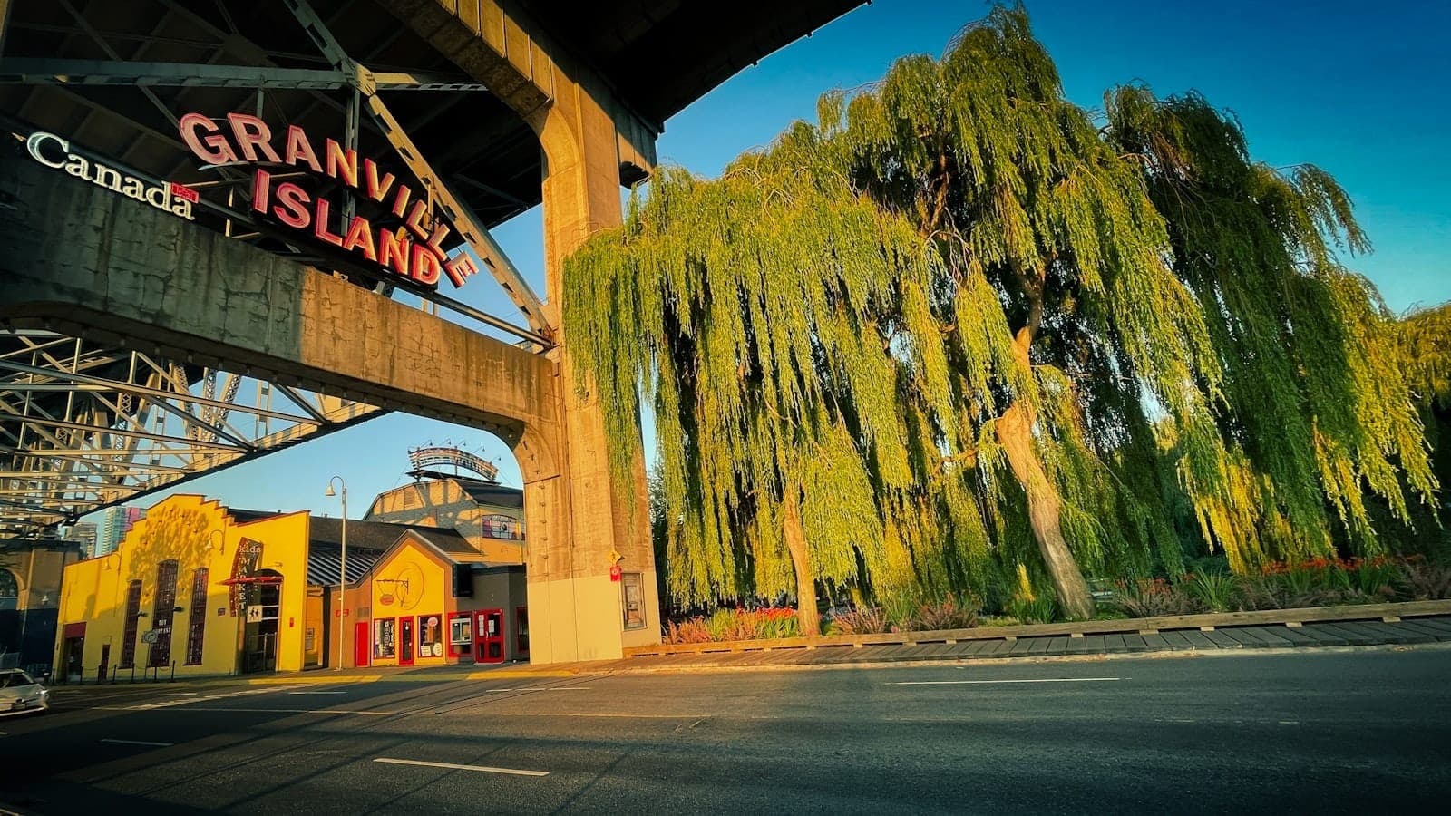 ป้ายทางเข้า Granville Island ใต้สะพาน ตึกสีเหลืองต้นไม้ใบเขียว แสงแดดสีทองสดใส