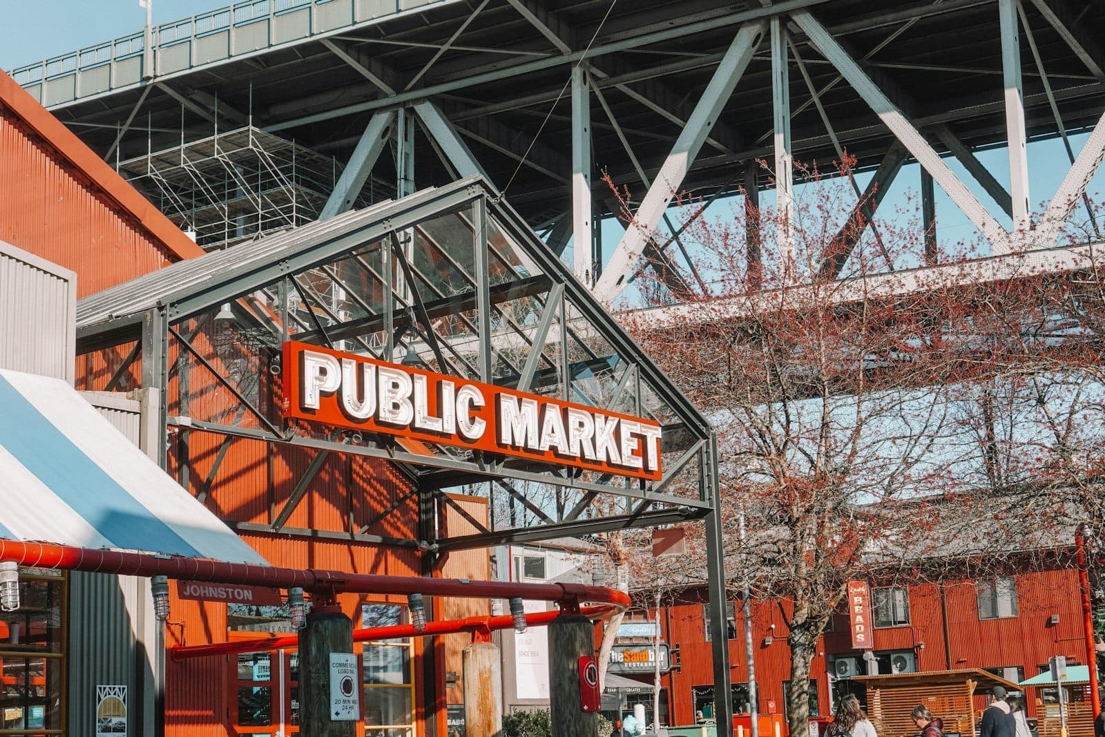 ทางเข้า Granville Island Public Market พร้อมป้ายสีแดงและขาวโดดเด่น โครงสร้างโลหะ และสะพานที่มองเห็นได้ด้านบนในวันที่แดดจ้า