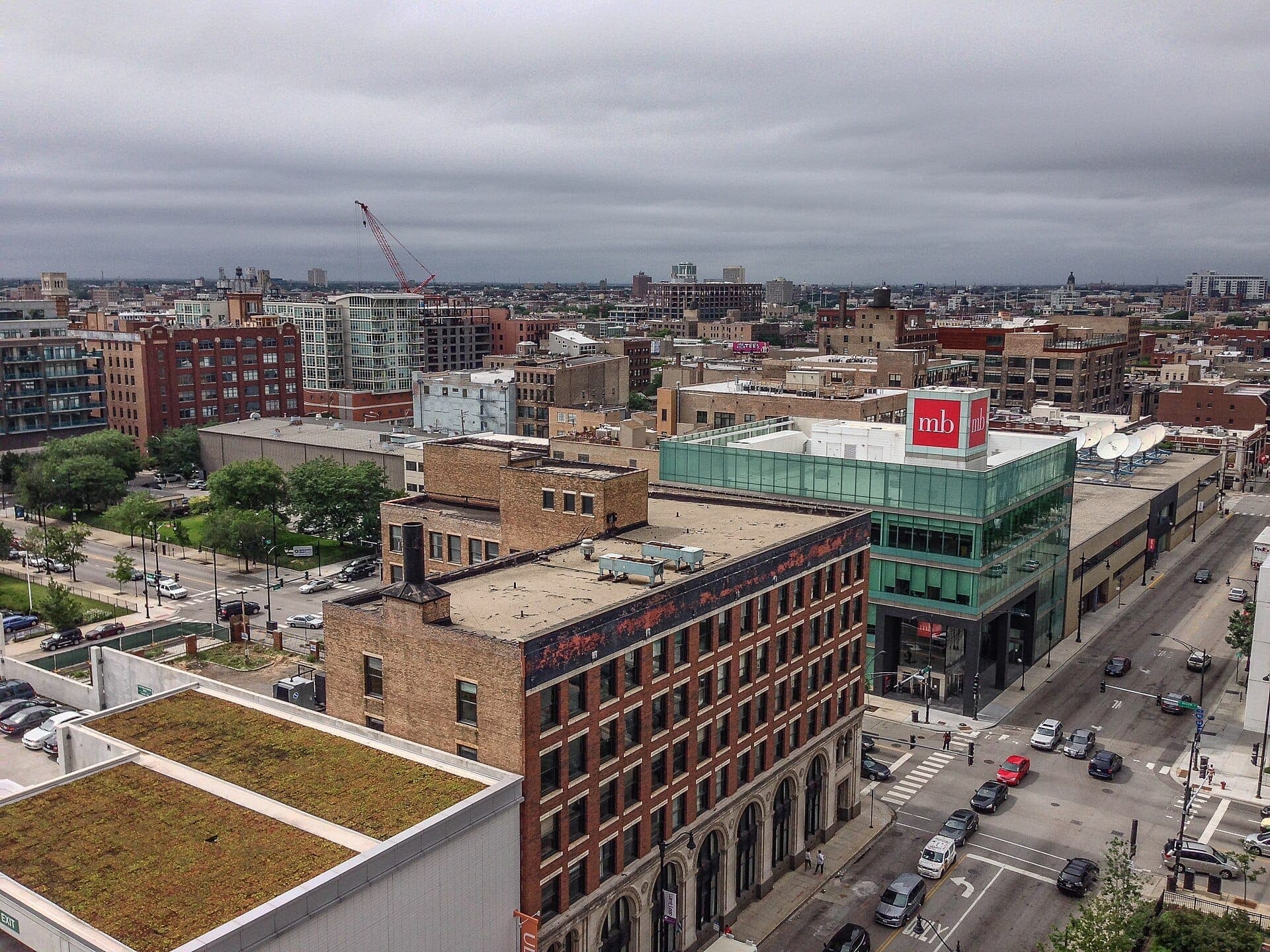 Aerial view of Chicago's Greektown and Halsted Street, showing brick buildings, modern glass structures, street traffic, and overcast skies.