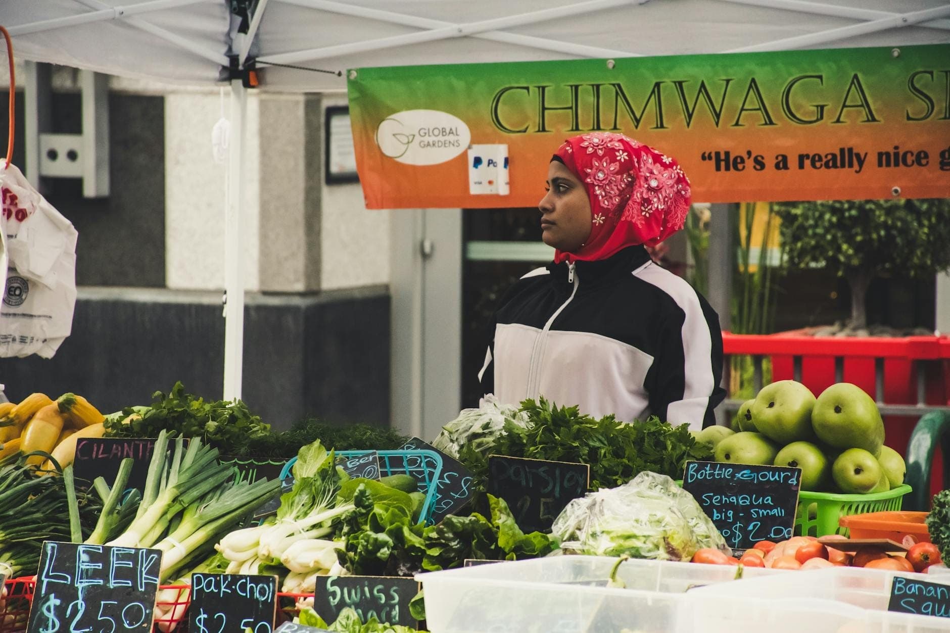 Farmer at a produce stand in an outdoor market, surrounded by fresh vegetables and handwritten price signs under a tent.