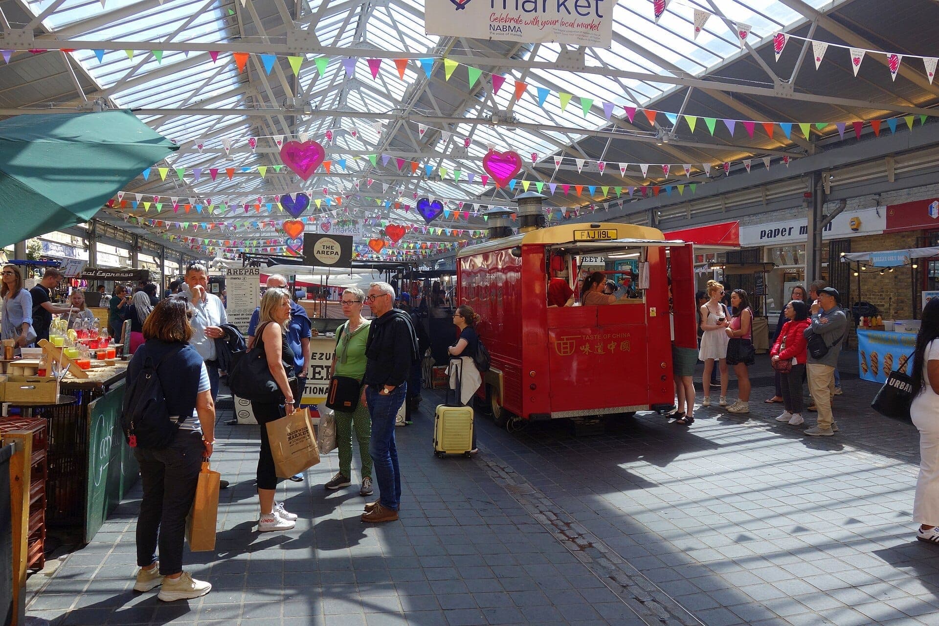 Besucher erkunden den belebten überdachten Greenwich Market mit bunten Wimpelketten, Essensständen und einem roten Vintage-Food-Truck unter einem Glasdach.