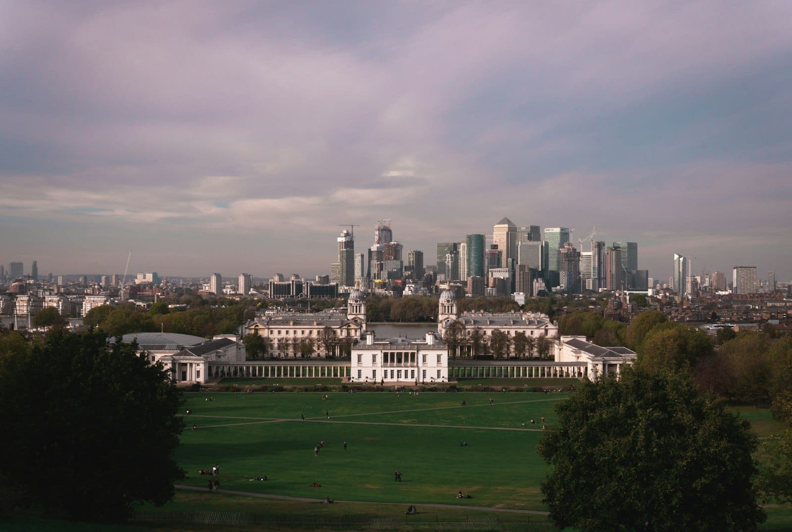Vue panoramique du parc de Greenwich, de la Queen's House et de l'Old Royal Naval College, avec la skyline moderne de Canary Wharf sous un ciel coloré au coucher du soleil.