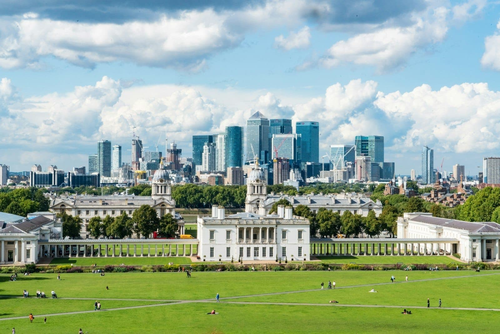 Blick auf die grünen Rasenflächen und prächtigen historischen Gebäude des Greenwich Park mit der modernen Skyline von Canary Wharf in London unter einem teilweise bewölkten Himmel.