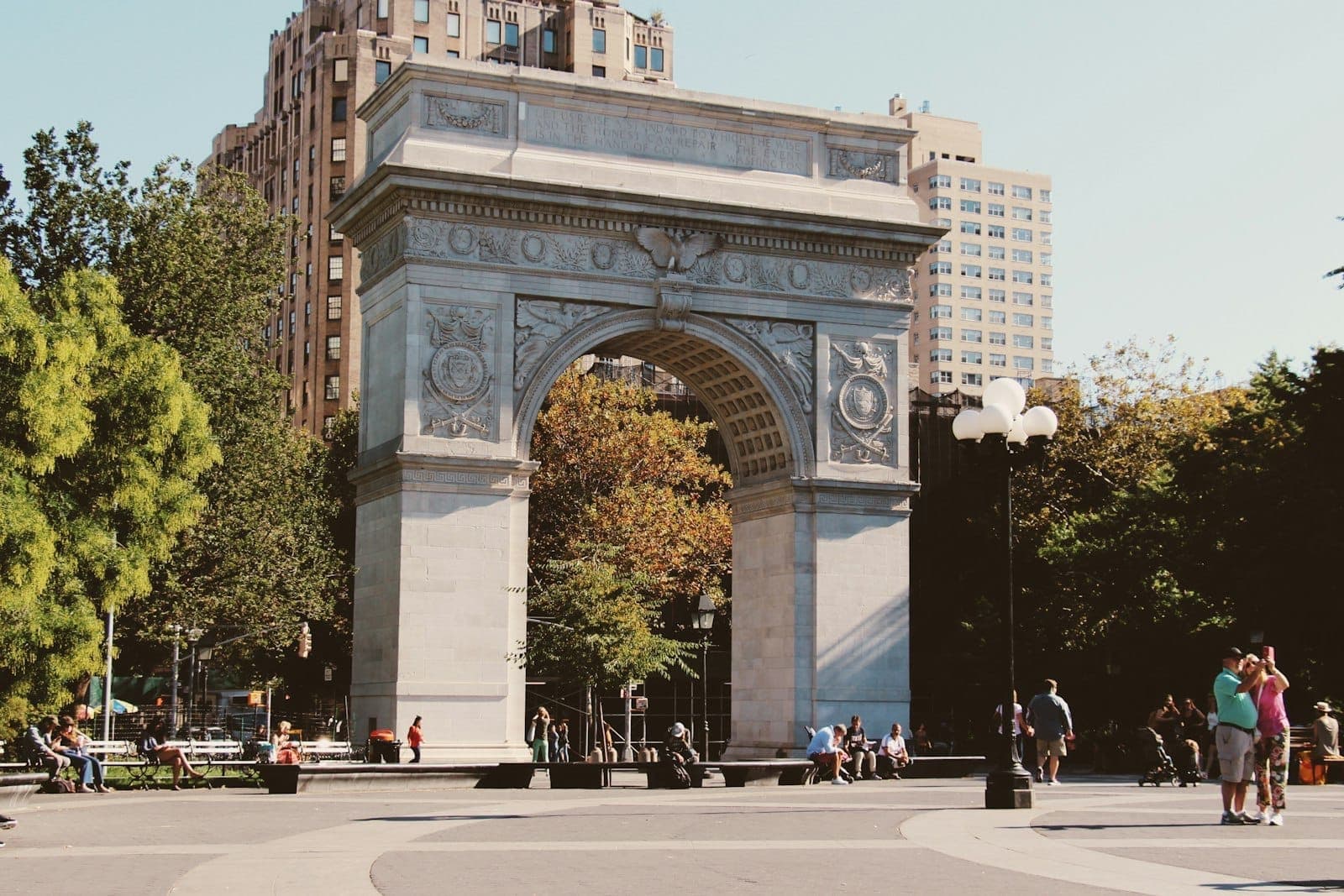 ภาพย่าน Washington Square Arch ใน Greenwich Village เวลาแดดจัด มีผู้คนเดินไปมา ต้นไม้และตึกนิวยอร์คคลาสสิกอยู่ด้านหลัง