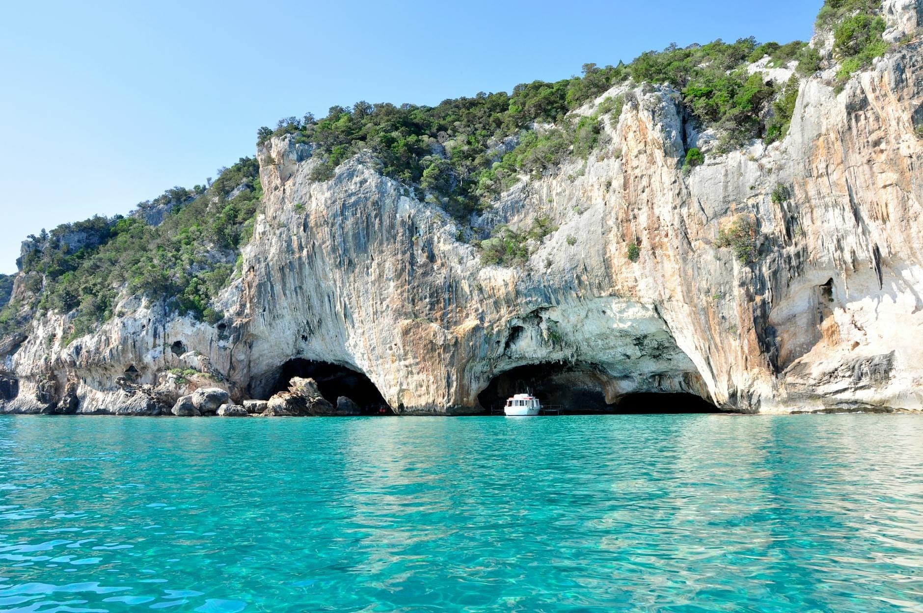 Vista de las entradas de la cueva marina Grotte del Bue Marino en los acantilados de piedra caliza, con agua turquesa y un barco cerca de la cueva bajo un cielo azul despejado.