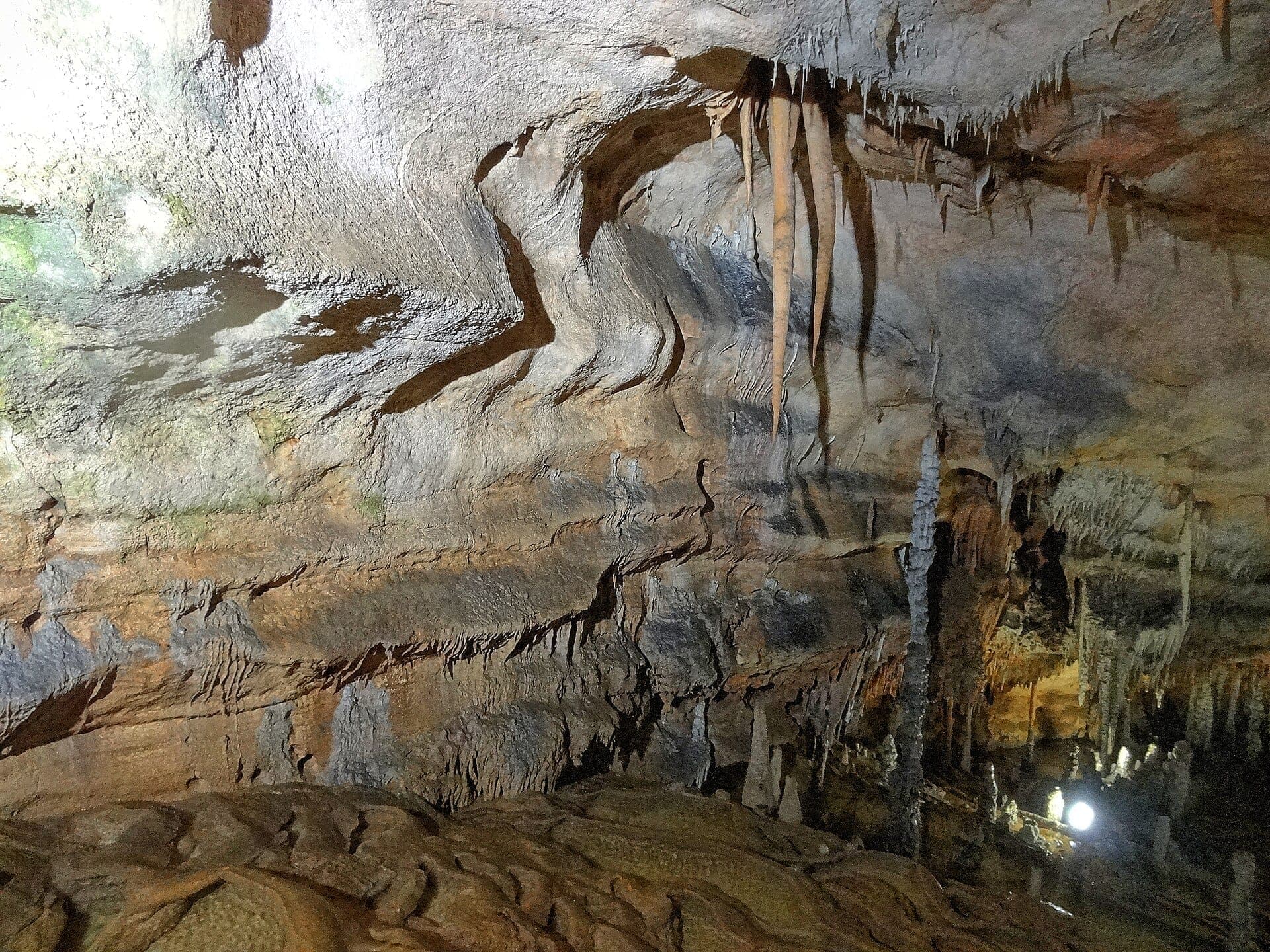 Estalactitas y formaciones de piedra caliza texturizadas dentro de la iluminada cueva marina Grotta del Fico en Baunei, Cerdeña, con colores naturales terrosos.