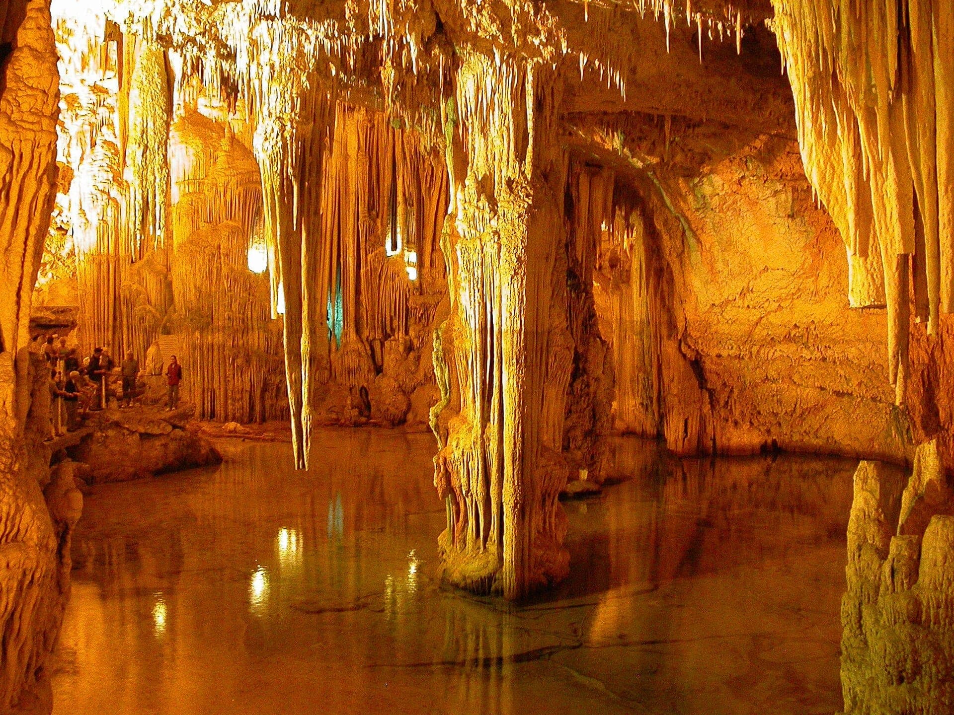 Visitantes caminan por un sendero dentro de las Grotte di Nettuno, rodeados de dramáticas estalactitas, estalagmitas y un reflectante lago subterráneo bajo una iluminación cálida.