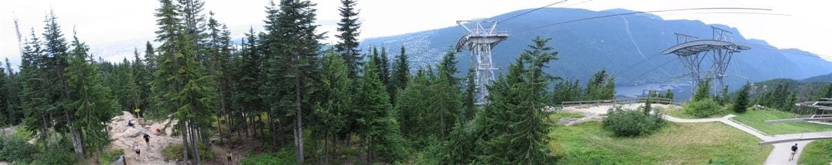 วิวพาโนรามาจากยอดเขา Grouse Grind ที่เห็นป่าสนทึบ พื้นที่โล่งหินขรุขระ เสากระเช้าไฟฟ้า และเทือกเขาในระยะไกลที่ Vancouver