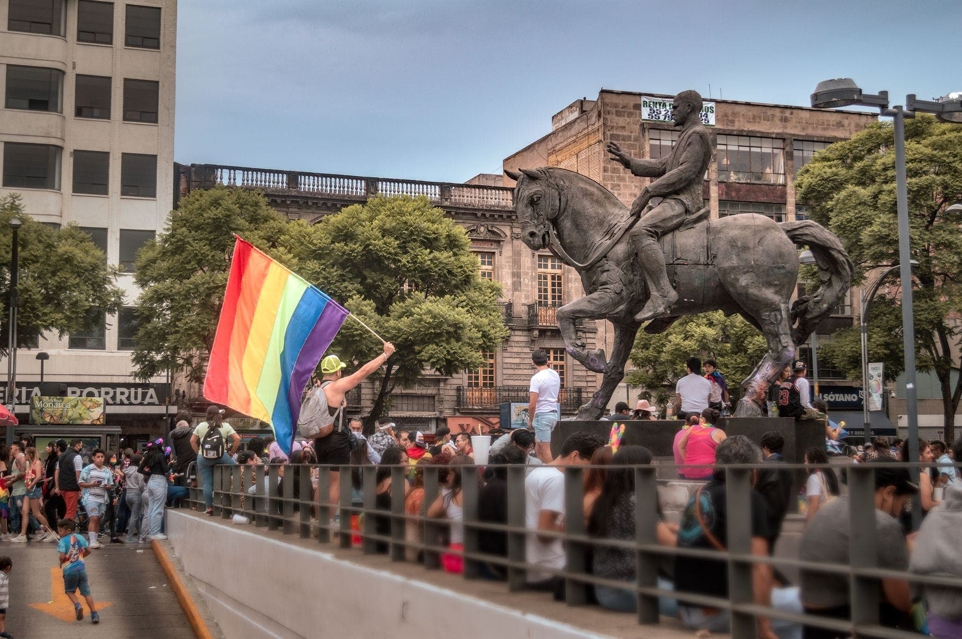 ฝูงชนรวมตัวกันรอบรูปปั้นขี่ม้าใจกลางเมือง Guadalajara ขณะที่มีคนหนึ่งโบกธงสีรุ้ง สะท้อนบรรยากาศอันคึกคักของงานสาธารณะ