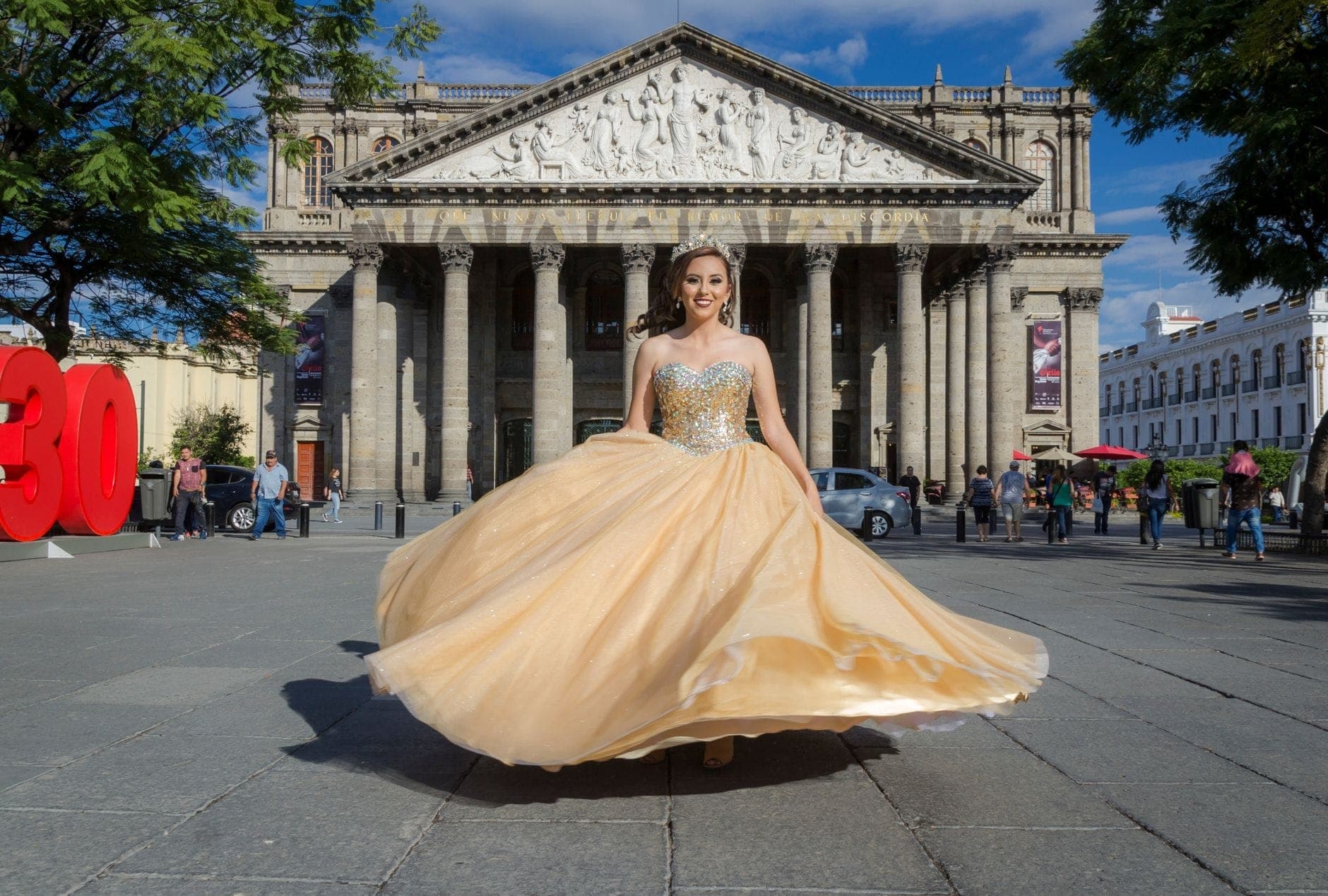 ผู้หญิงในชุดทางการหมุนตัวอยู่หน้า Teatro Degollado สถาปัตยกรรมนีโอคลาสสิกอันเป็นสัญลักษณ์ของกัวดาลาฮารา โดยมีผู้คนและป้ายแบนเนอร์ปรากฏอยู่ในลานกว้าง