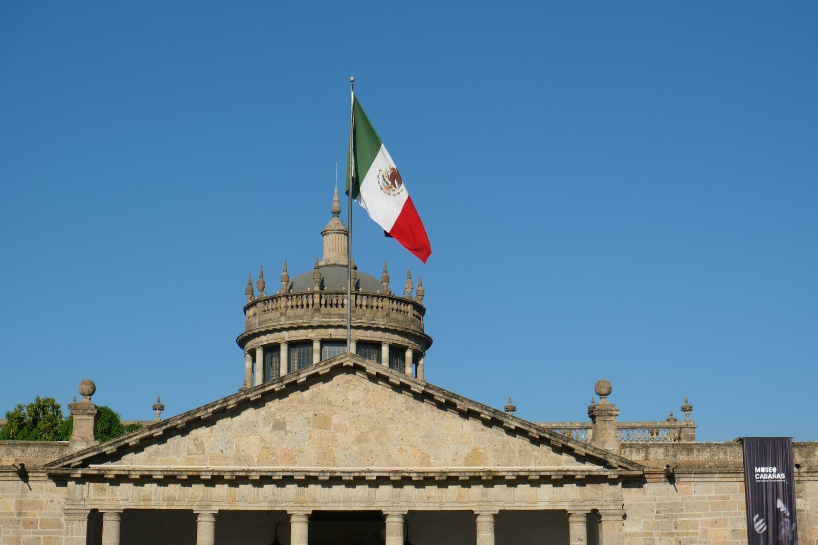 Hospicio Cabañas in Guadalajara with a large Mexican flag and clear blue sky, showing iconic neoclassical architecture.