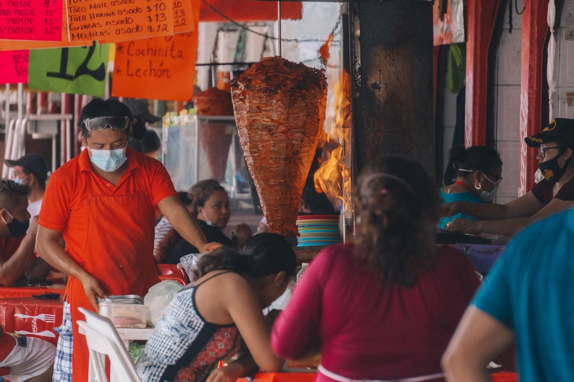 แผงอาหารริมถนนที่คึกคักในกัวดาลาฮาราพร้อมผู้คนที่กำลังรับประทานอาหาร พ่อครัวที่กำลังเสิร์ฟ และทรอมโปขนาดใหญ่ที่มีเนื้อย่างบนเปลวไฟ