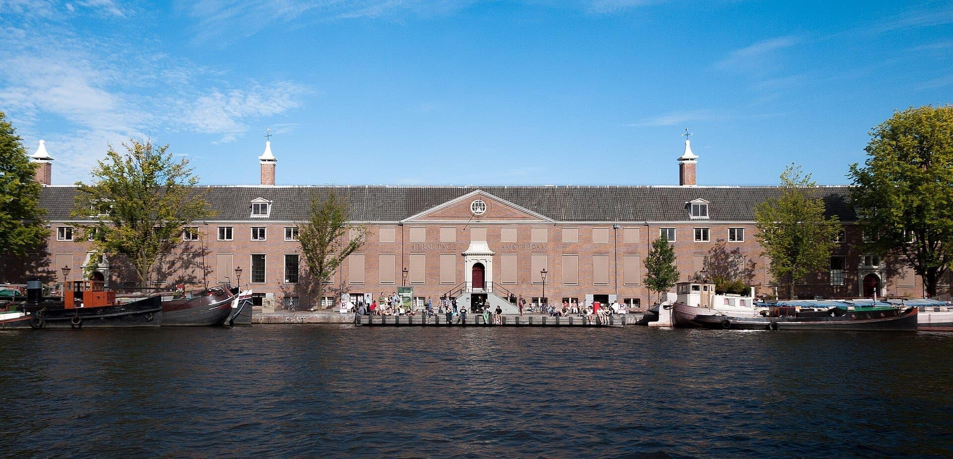 Das H'ART Museum Amsterdam vom anderen Ufer der Amstel aus gesehen, mit seiner historischen Backsteinfassade, davor vertäuten Booten und eintretenden Besuchern.