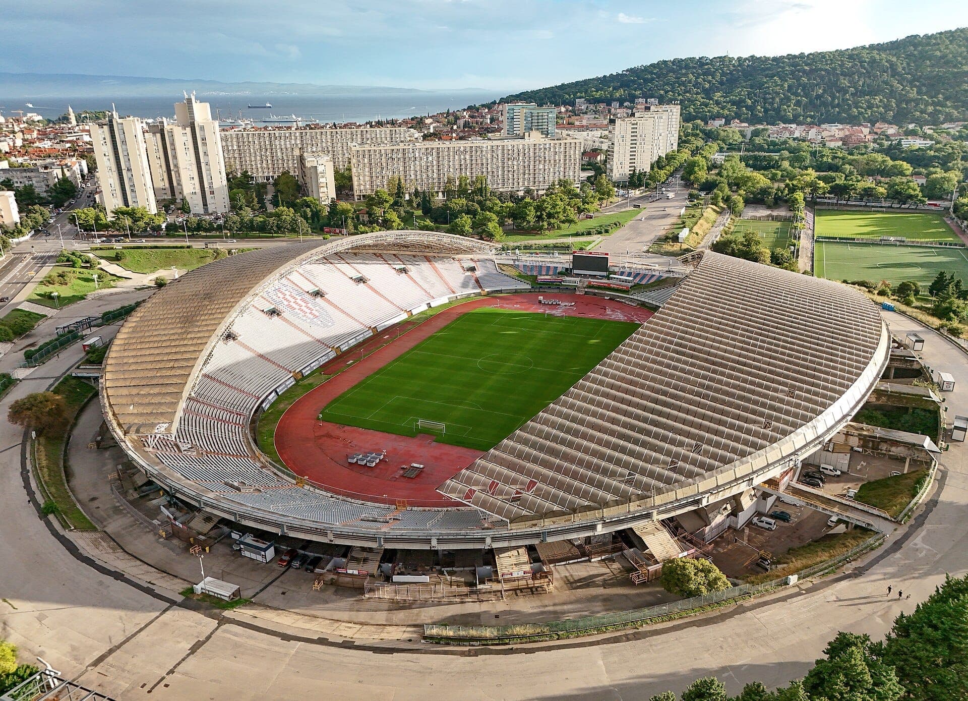 Vista aerea dello Stadio Poljud a Spalato, in Croazia, con la sua struttura ovale, il campo di calcio verde, i palazzi circostanti e le colline vicine alla luce del giorno.