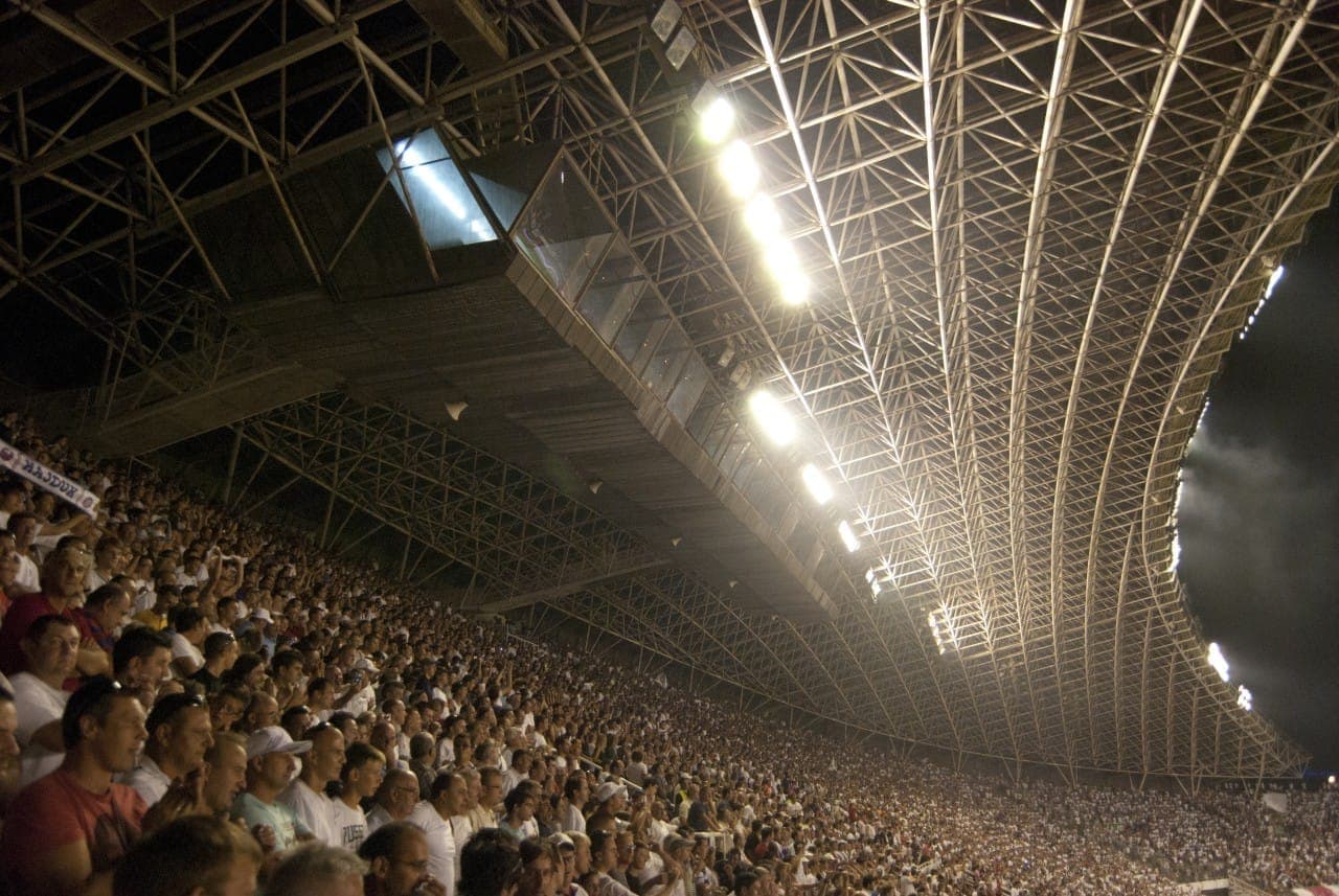 Tribune affollate allo Stadio Poljud, Split, piene di tifosi di calcio sotto il caratteristico tetto in acciaio curvilineo e le luci brillanti dello stadio.