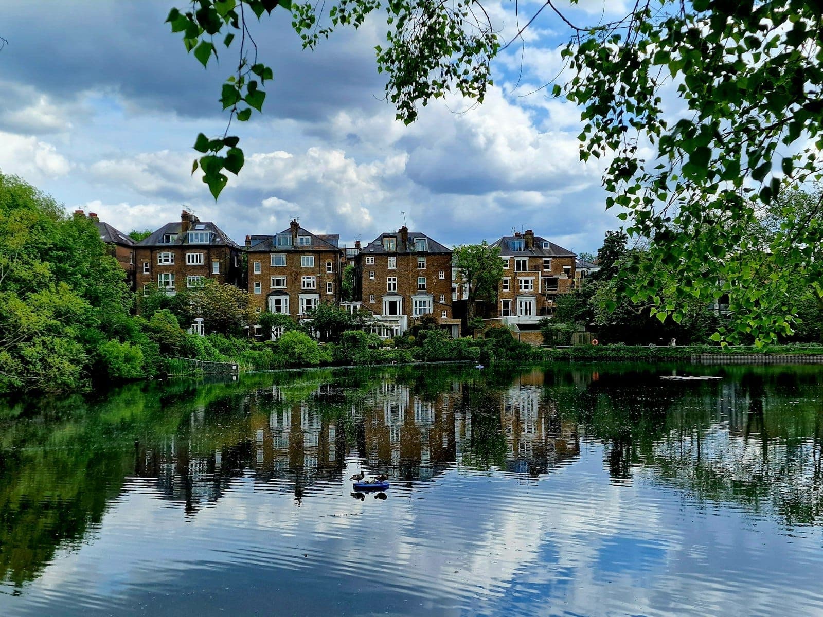 Maisons en briques pittoresques reflétées dans un étang de Hampstead Heath, encadrées par une végétation luxuriante et un ciel dramatique, capturant l'essence de Hampstead & Highgate.