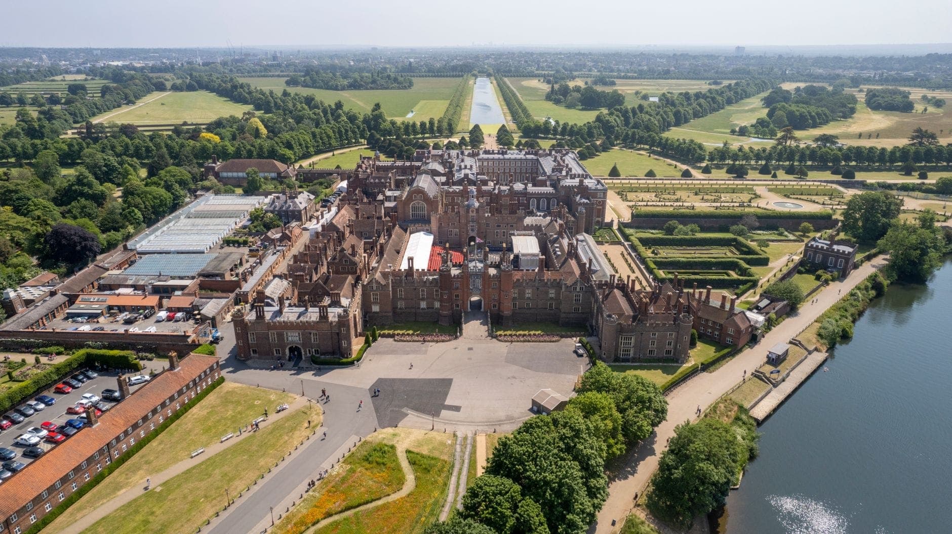 Ein atemberaubender Luftblick auf Hampton Court Palace, umgeben von üppigen Gärten, der Themse und weitläufigen Parkanlagen unter klarem Himmel.