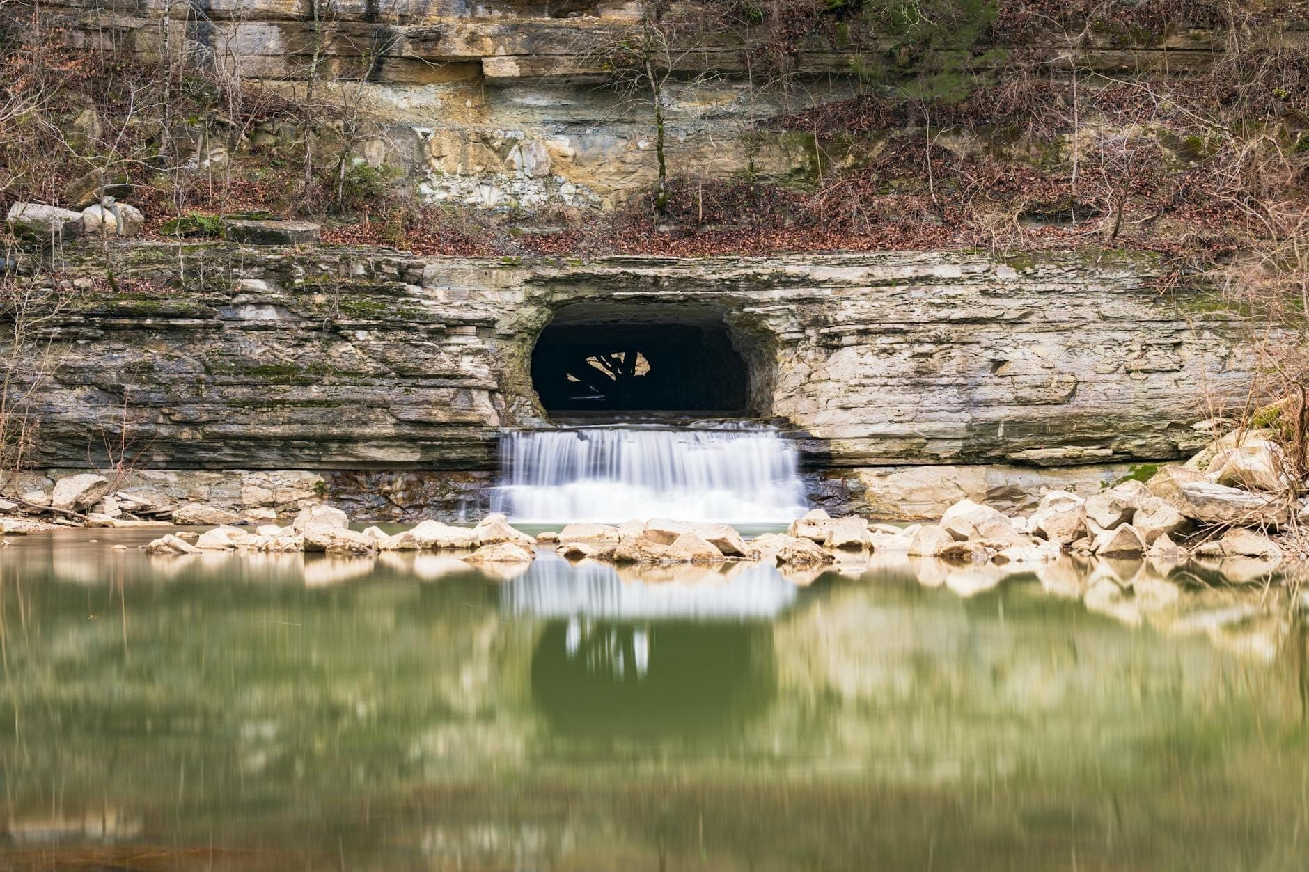 A hand-dug stone tunnel with water gently flowing out into a calm river, surrounded by rocky banks and natural cliff walls.