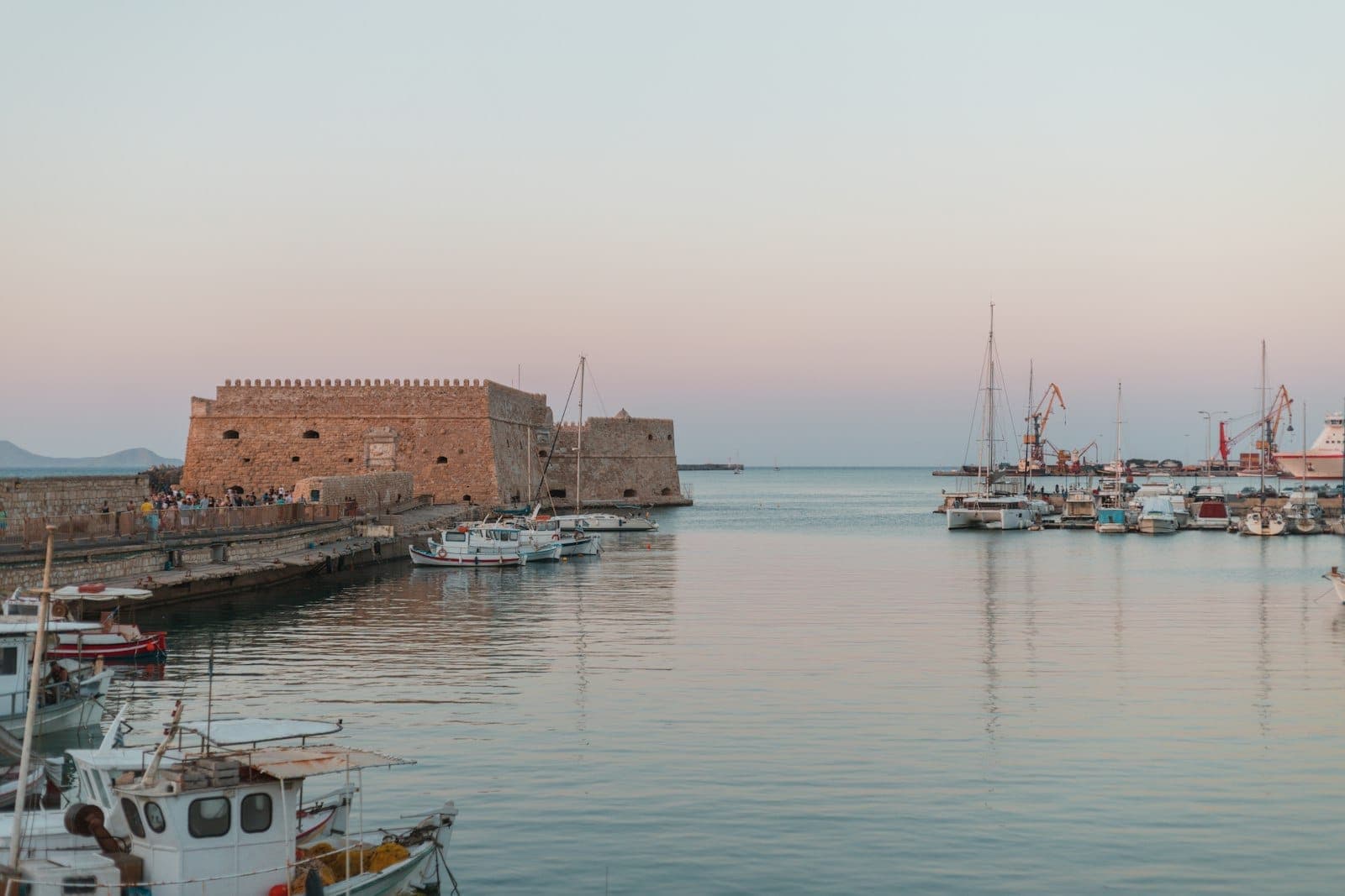 Blick auf den Hafen von Heraklion bei Sonnenuntergang mit der venezianischen Festung auf der linken Seite, ruhigem Wasser, Booten und einem pastellfarbenen Himmel.