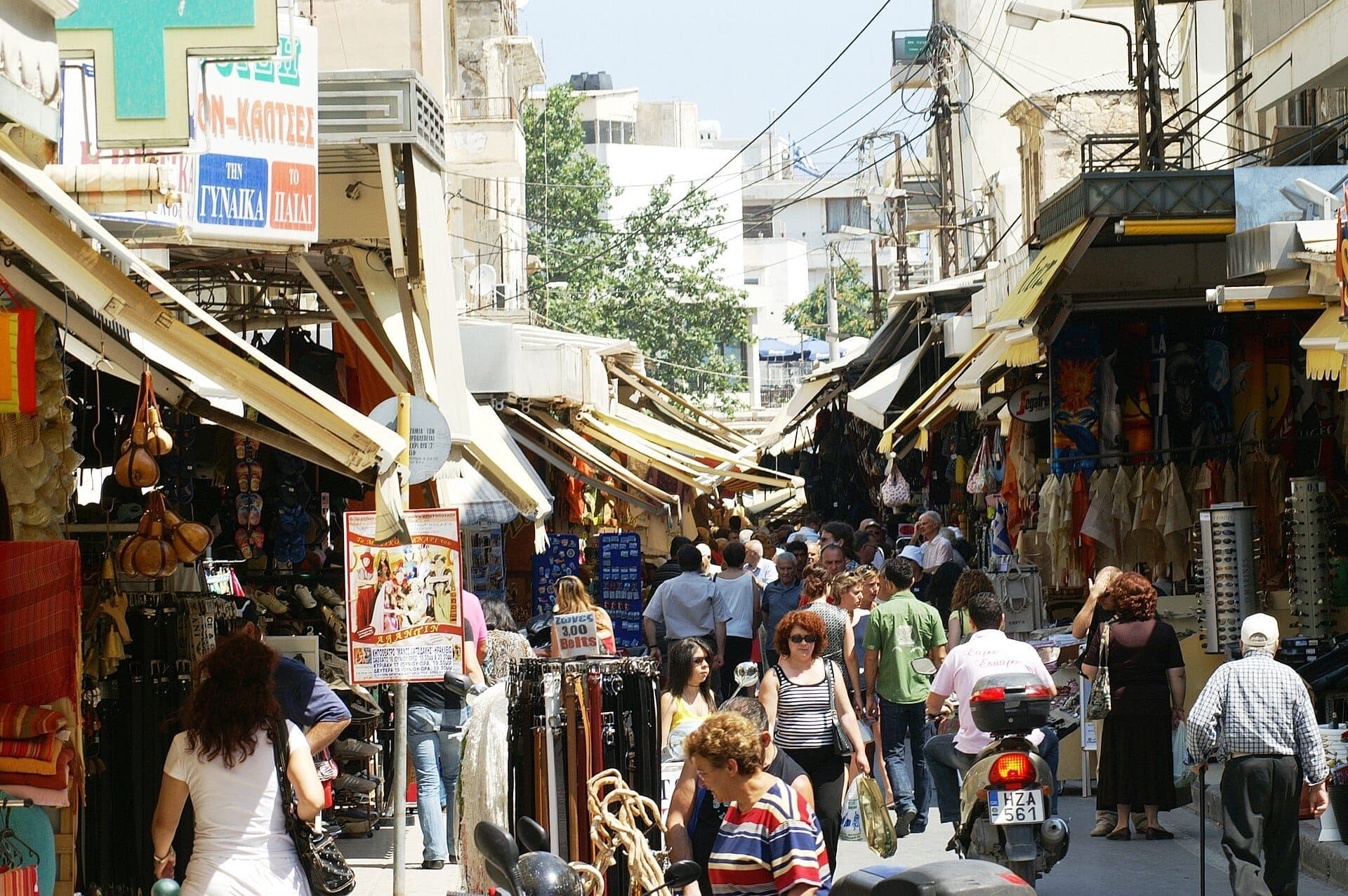 Belebte Tagesszene auf dem Markt der Straße 1866 in Heraklion, mit Menschen, die zwischen offenen Ladengeschäften, Markisen und bunten Marktständen schlendern.