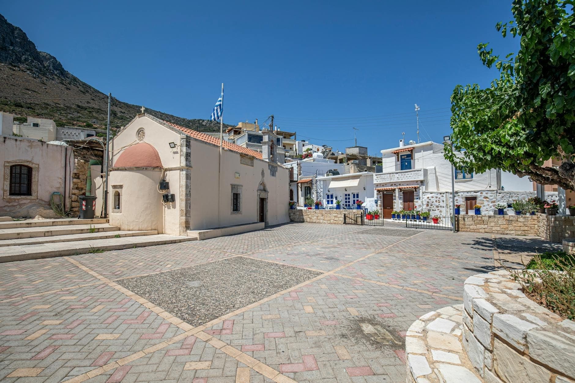 Ein charmanter Dorfplatz auf Kreta mit einer traditionellen Kirche, weiß getünchten Häusern und griechischer Flagge an einem sonnigen Tag.
