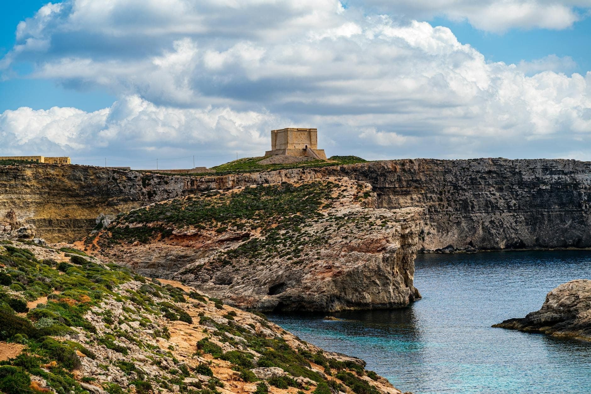 Dramatische Küstenklippen und Buchten in Malta mit einem historischen Turm auf dem Hügel, unter teilweise bewölktem Himmel, einladend für Naturwanderungen.