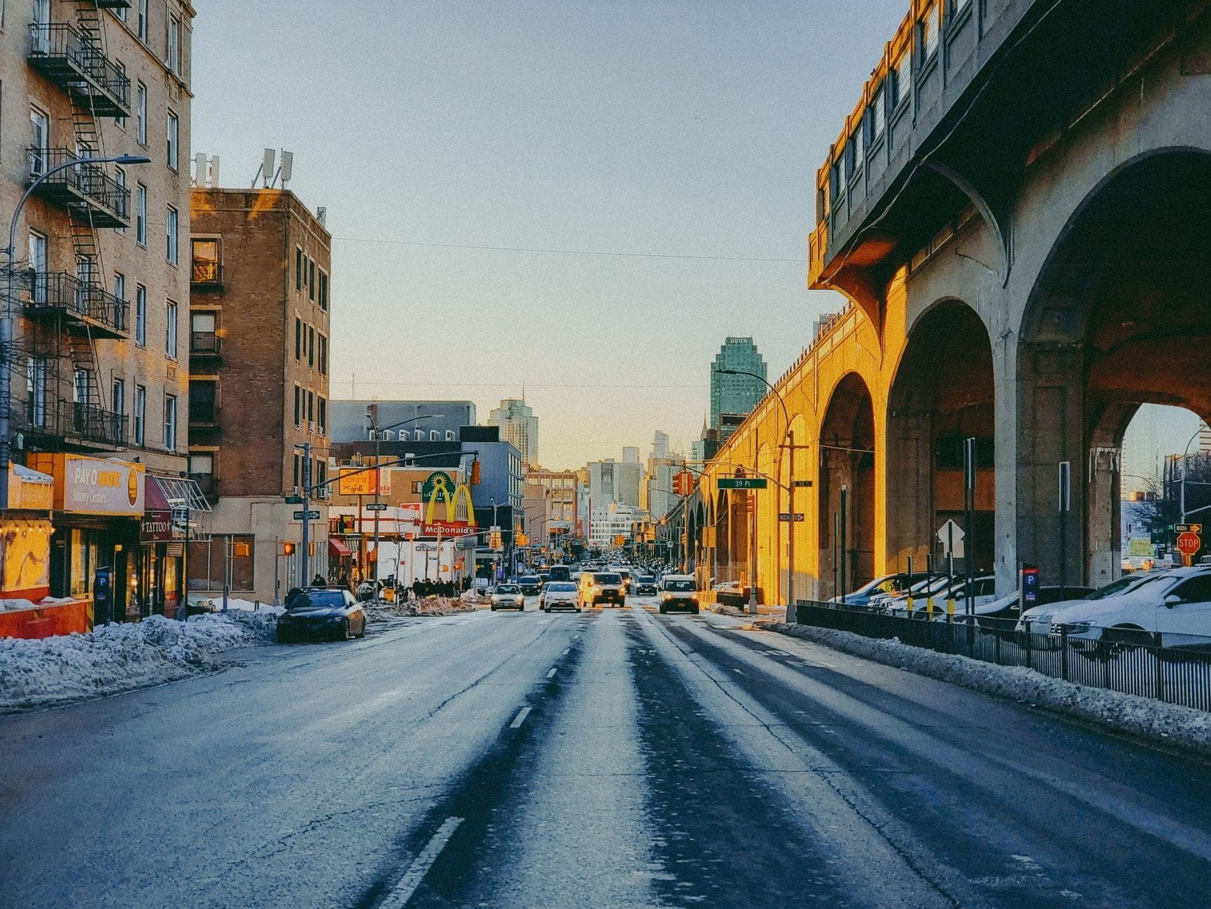 ถนนในเมืองที่ Queens หรือ Brooklyn พร้อมรางรถไฟใต้ดินยกระดับ อาคารวินเทจ และเส้นขอบฟ้าที่อยู่ไกลออกไป ภายใต้แสงอาทิตย์สีทองในฤดูหนาว