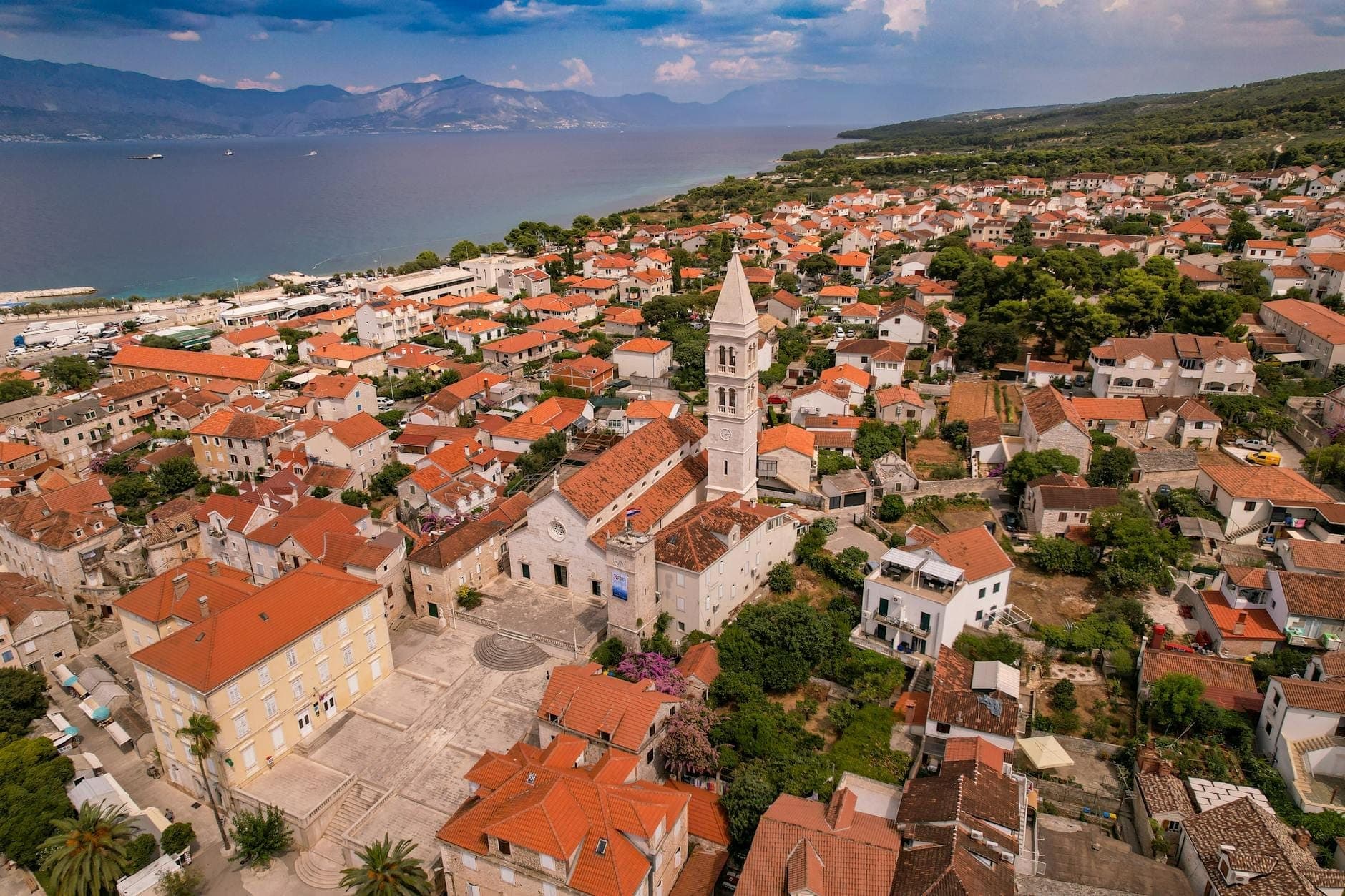 Vista aerea di una piccola cittadina costiera vicino a Spalato, con una chiesa dal alto campanile, case dai tetti rossi e il mare blu sullo sfondo.