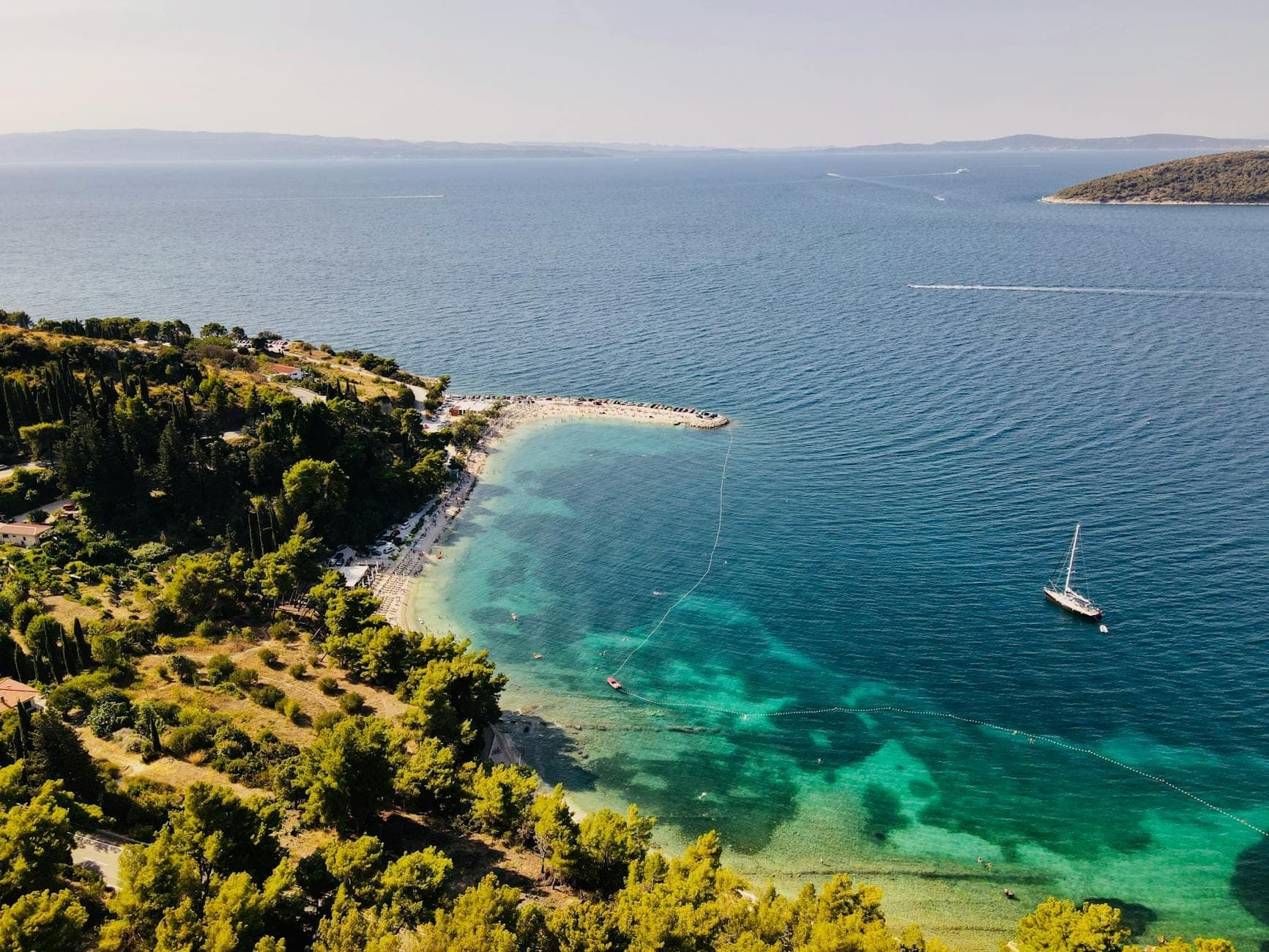 Vista aerea di una tranquilla spiaggia rocciosa vicino a Spalato con acqua turchese, riva boscosa e uno yacht ancorato al largo sotto un cielo sereno.