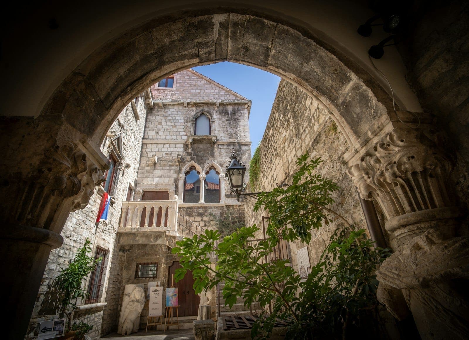 Cortile medievale in pietra a Spalato, incorniciato da un antico arco, con rigogliosa vegetazione, colonne ornate e la luce del sole sulle facciate storiche.