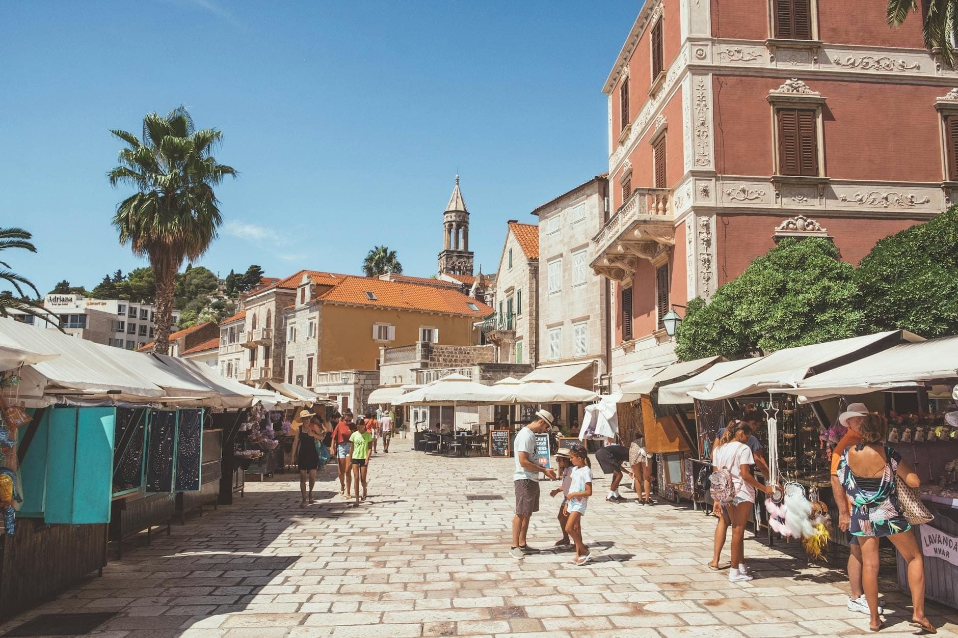 Scena di mercato all'aperto a Spalato con persone che fanno acquisti alle bancarelle lungo una piazza lastricata di pietra circondata da storici edifici mediterranei.
