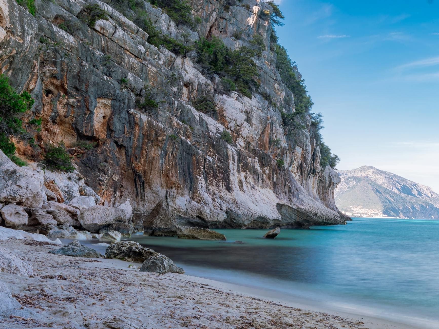 Acantilados rocosos y playa de arena recóndita bordeada de agua turquesa, que sugiere una playa de acceso a pie típica del pintoresco litoral de Cerdeña.