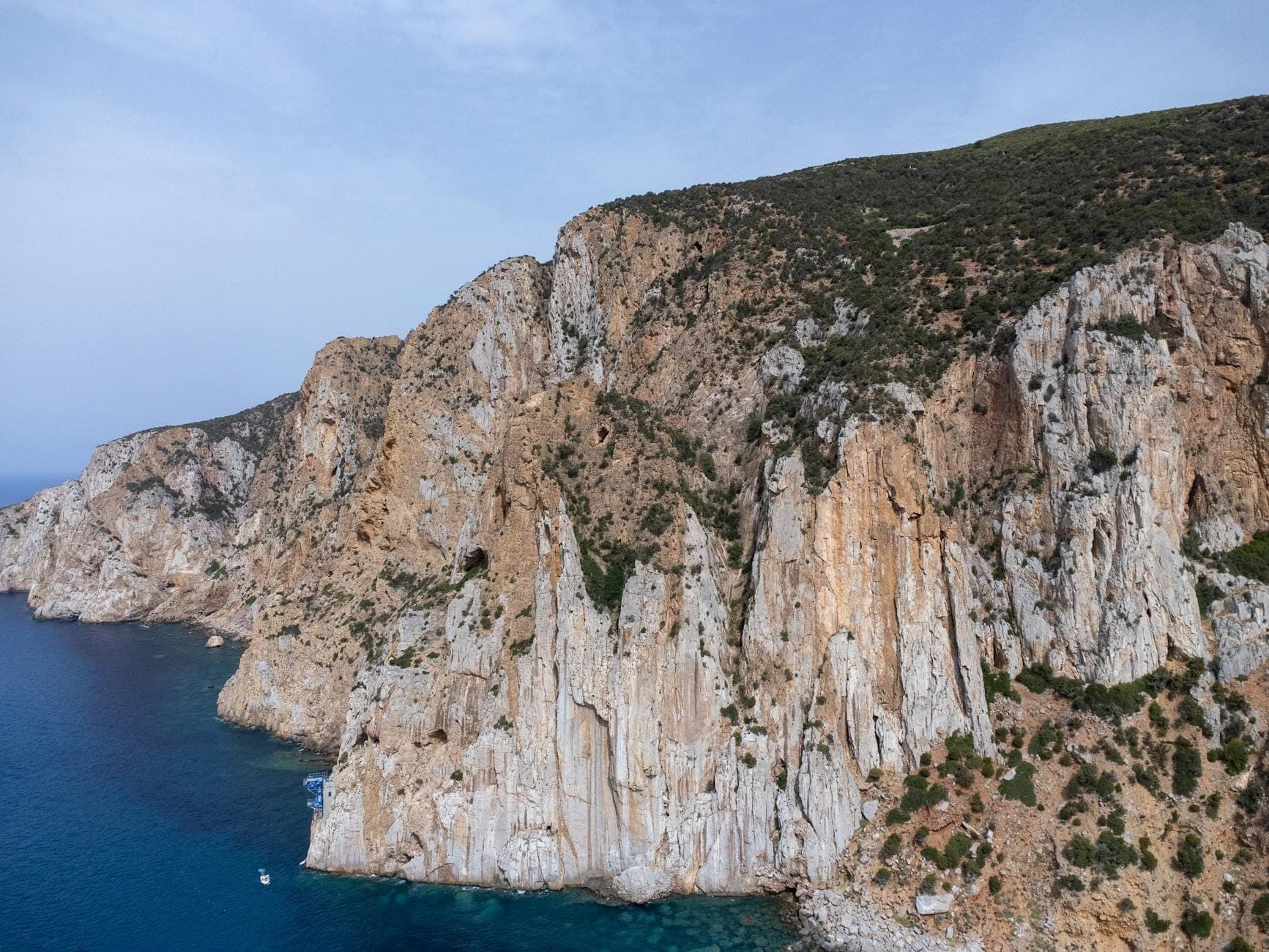 Escarpados acantilados que se elevan sobre el mar azul profundo, con vegetación escasa en terreno rocoso inclinado bajo un cielo despejado.