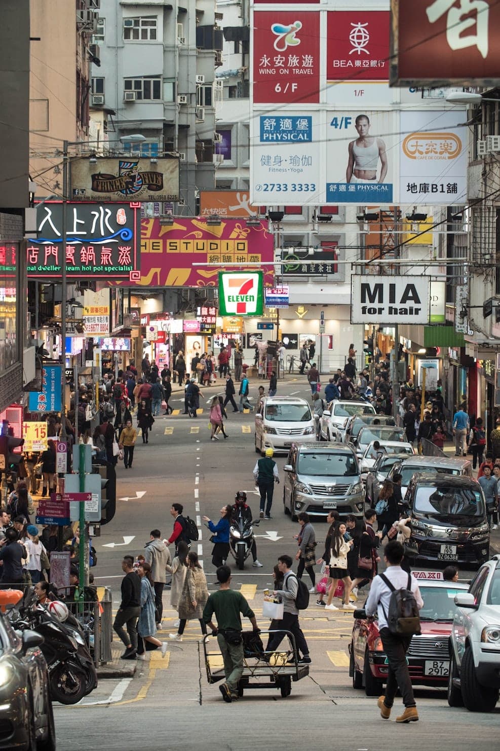 Crowded street in Hong Kong with heavy pedestrian traffic, cars, neon signs, and dense urban buildings