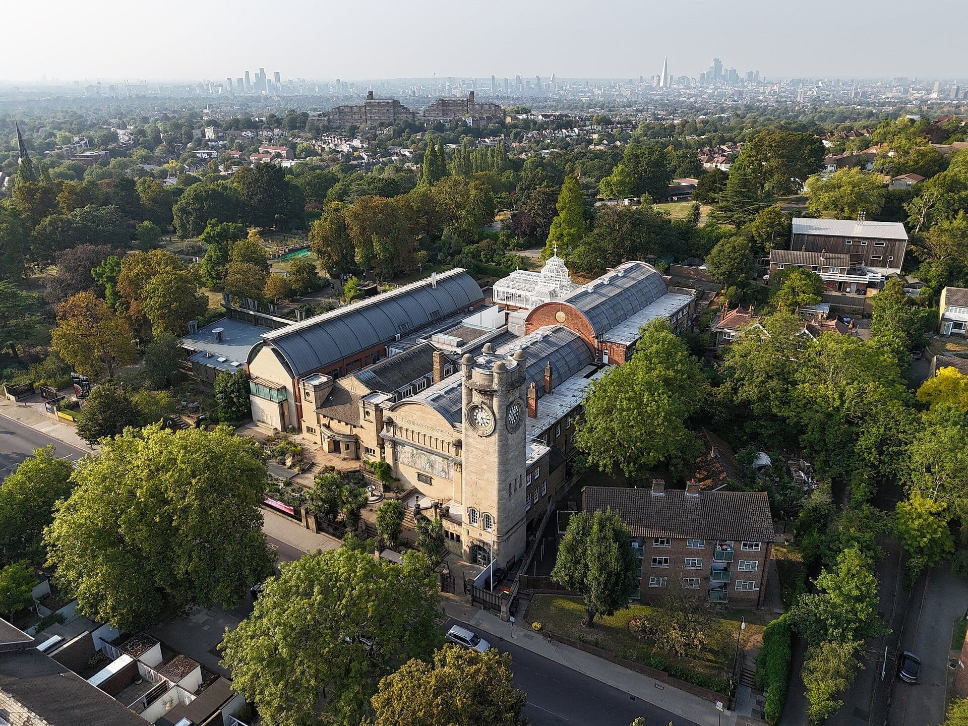 Luftaufnahme des Horniman Museum and Gardens, umgeben von üppigem Grün, mit der Skyline von Zentral-London in der dunstigen Ferne.