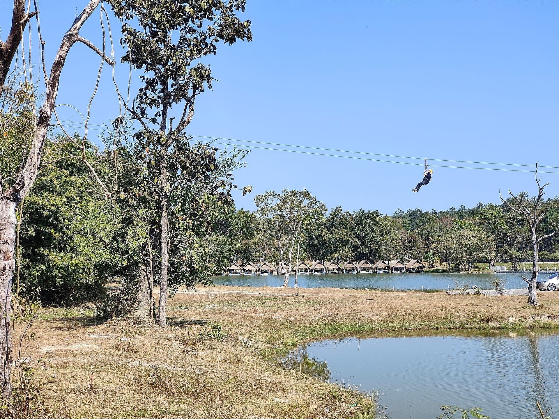 Eine Person beim Zipline über den Huay Tung Tao See, mit Bambushütten am Ufer, Bäumen und blauem Himmel im Mittagslicht.