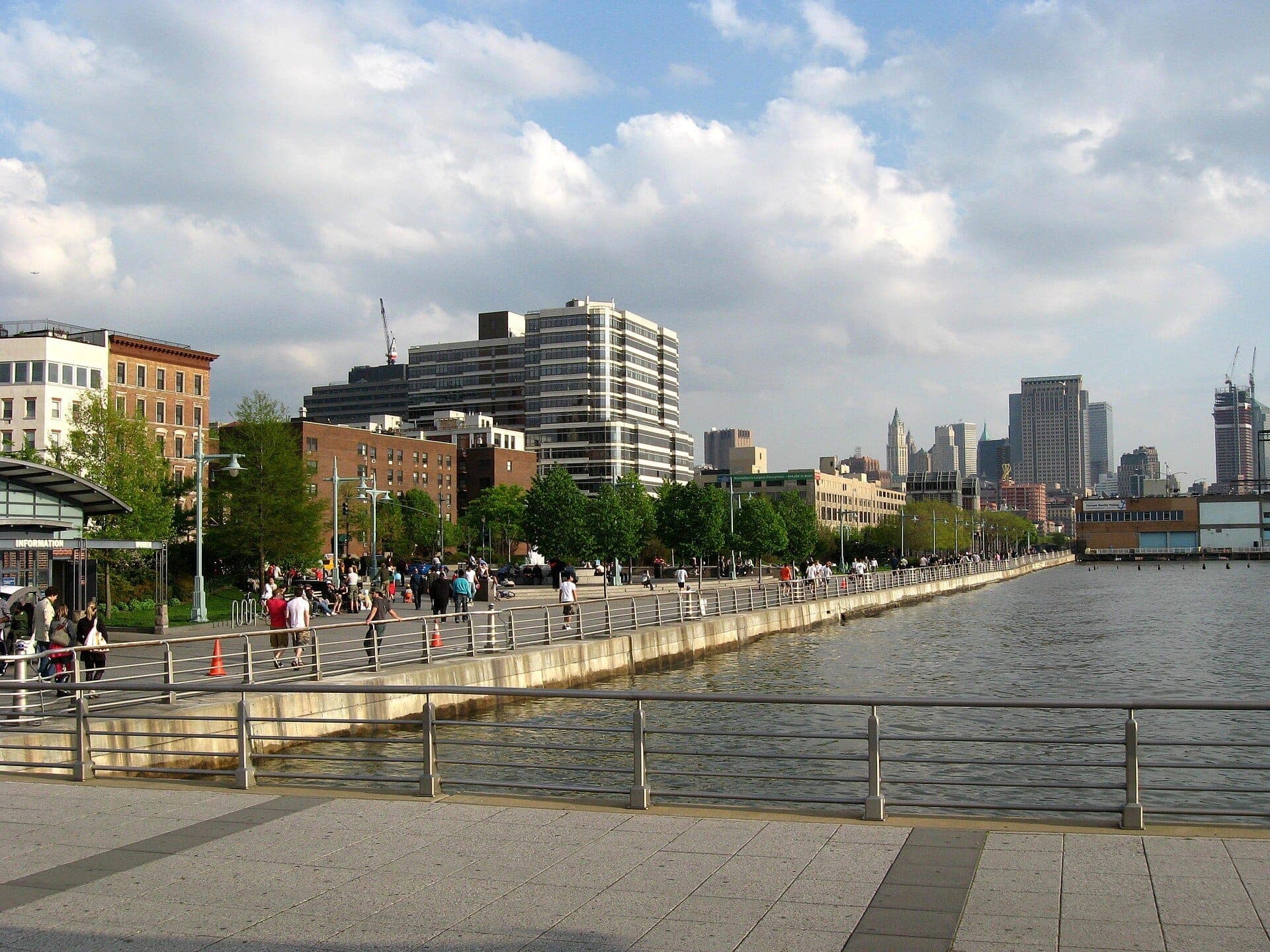 ผู้คนเดินเล่นตามทางเดินริมน้ำของ Hudson River Park ท่ามกลางต้นไม้ สายน้ำ และตึกระฟ้าของ Manhattan ใต้ท้องฟ้ามีเมฆบางส่วน