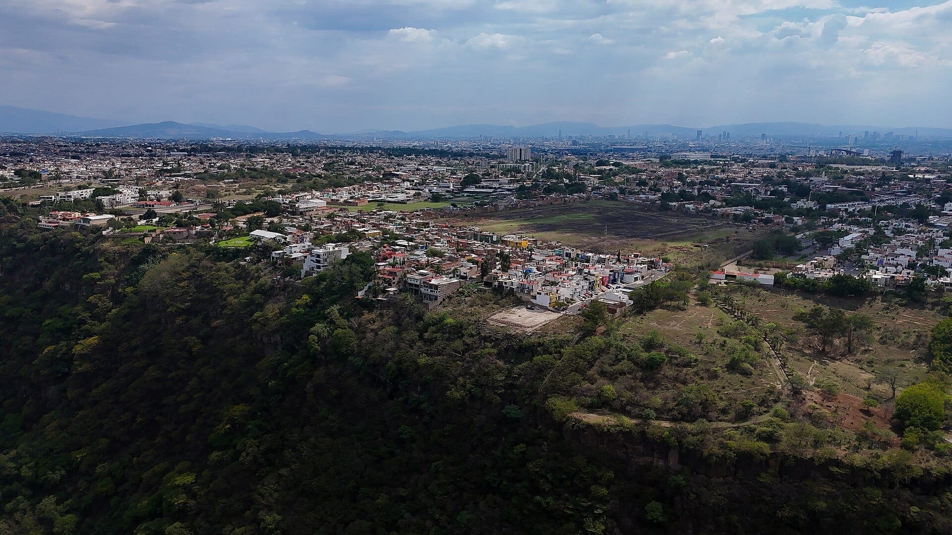 มุมมองทางอากาศของย่าน Huentitán และหน้าผา พร้อมด้วยหุบเขา Barranca de Oblatos และเมือง Guadalajara ที่กว้างใหญ่เป็นฉากหลังใต้ท้องฟ้าที่มีเมฆปกคลุม