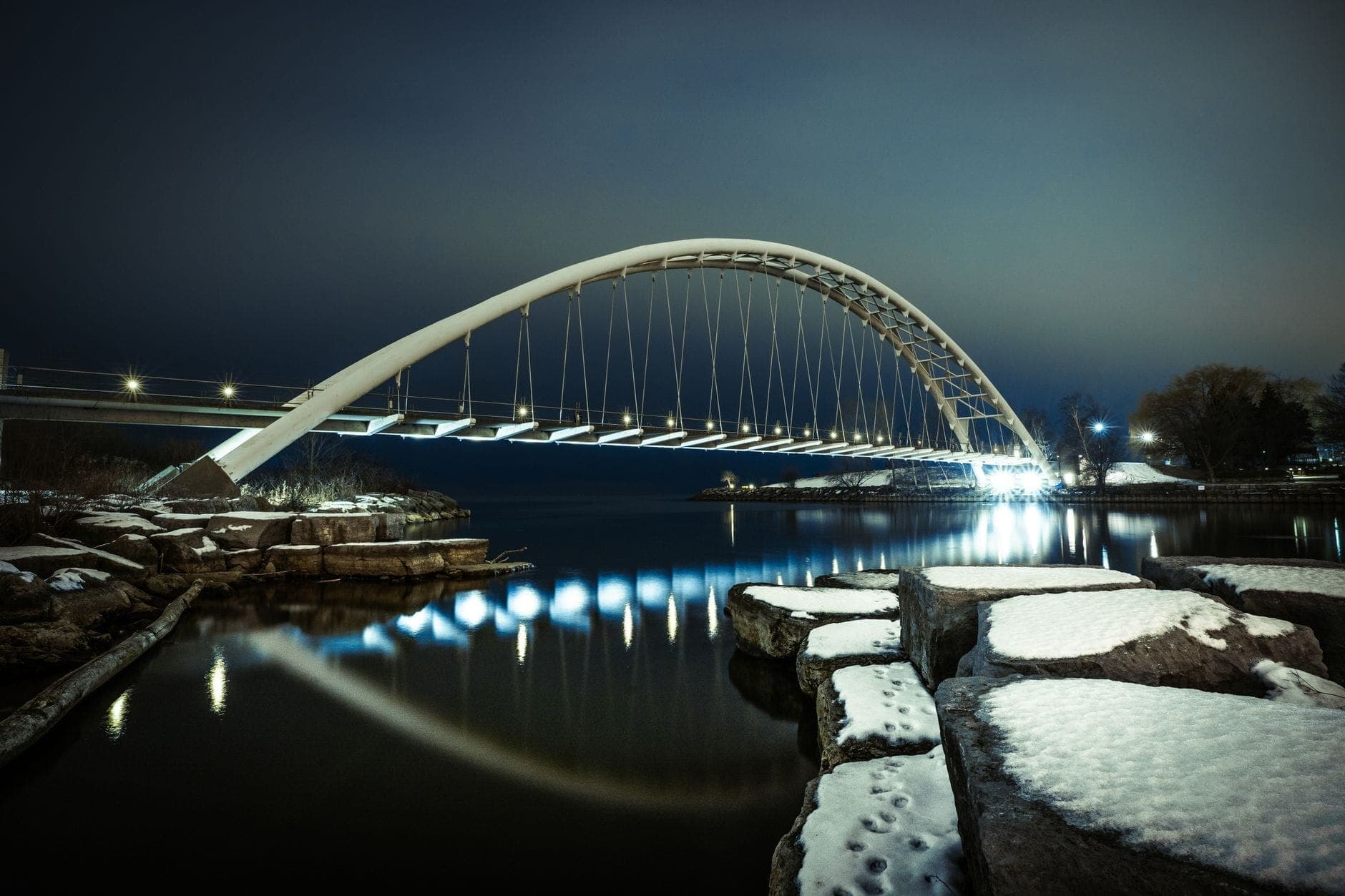 Vista notturna del Humber Bay Arch Bridge illuminato, con il suo riflesso nelle acque calme del fiume Humber e la neve sulle rocce vicine.
