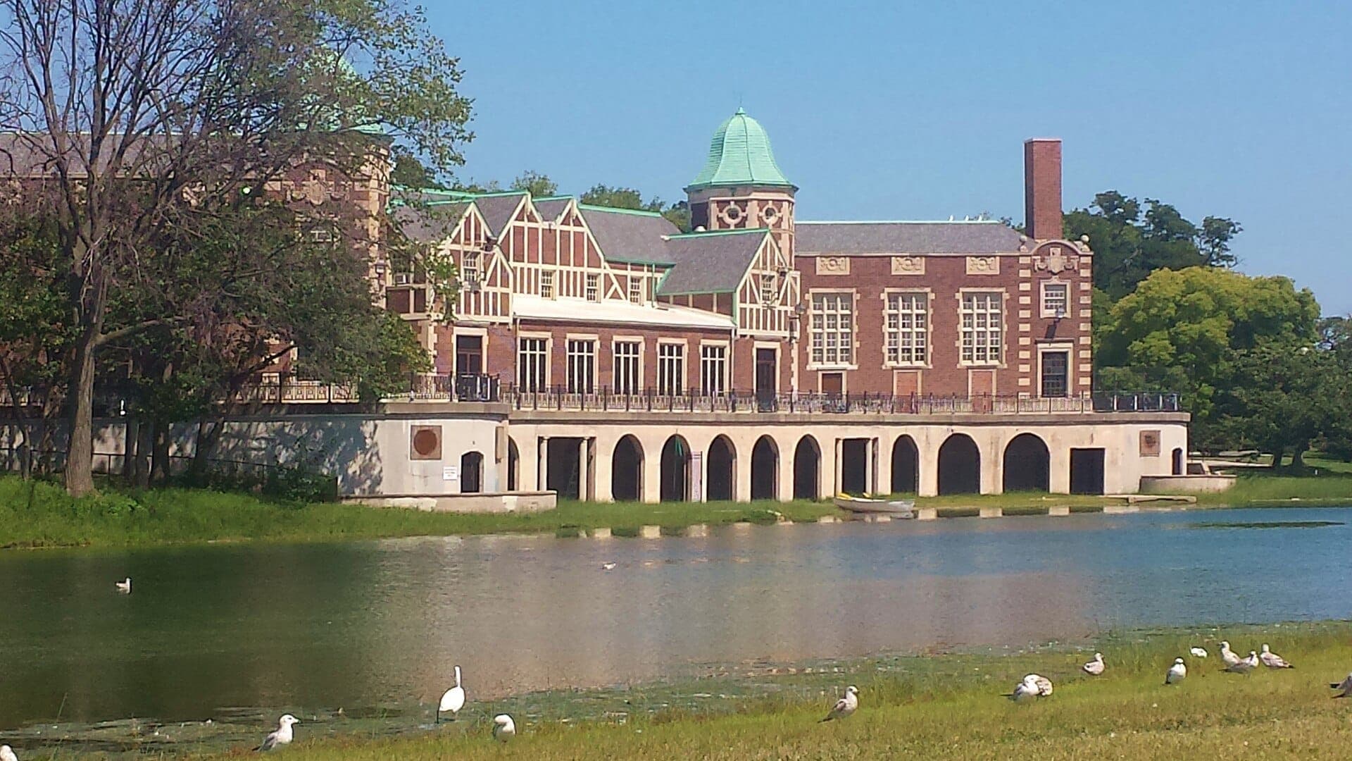 Historic Humboldt Park boathouse with arched colonnade overlooks a calm lagoon, surrounded by greenery and birds on a sunny day.