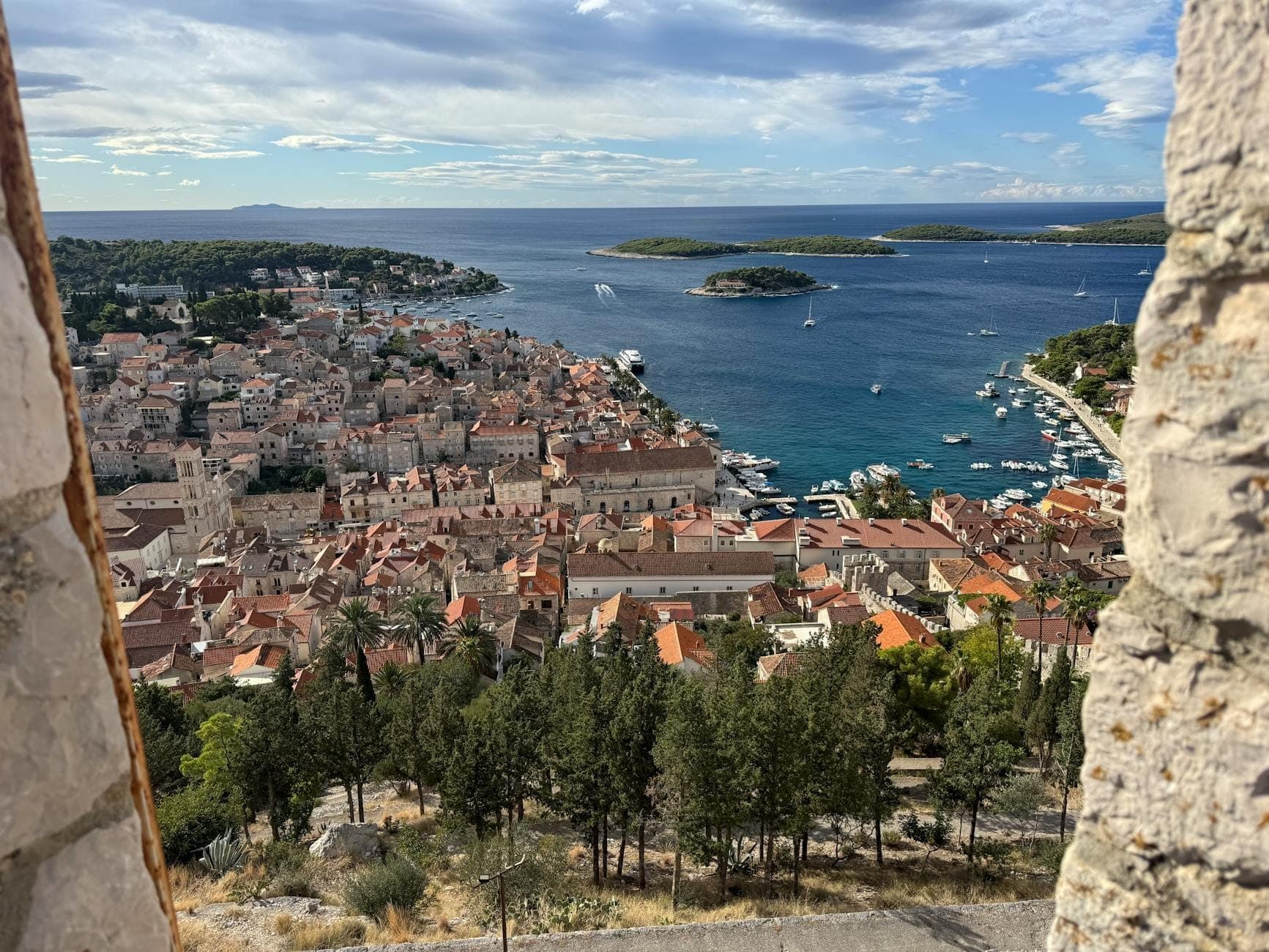 Vue panoramique de la ville et du port de Hvar depuis les hauteurs, avec ses toits rouge brique, la mer bleue parsemée de bateaux, les pins verts et les îles environnantes sous un ciel partiellement nuageux.