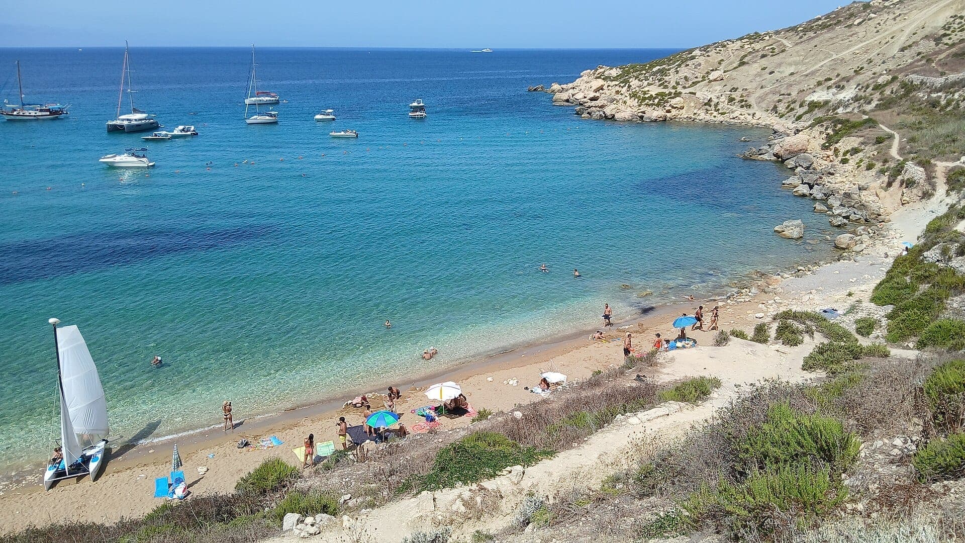 Ein Sandstreifen der Imgiebah Bay mit türkisfarbenem Wasser, kleinen vor Anker liegenden Booten, Sonnenbadenden, Sonnenschirmen und trockenen Felsenklippen im Hintergrund.